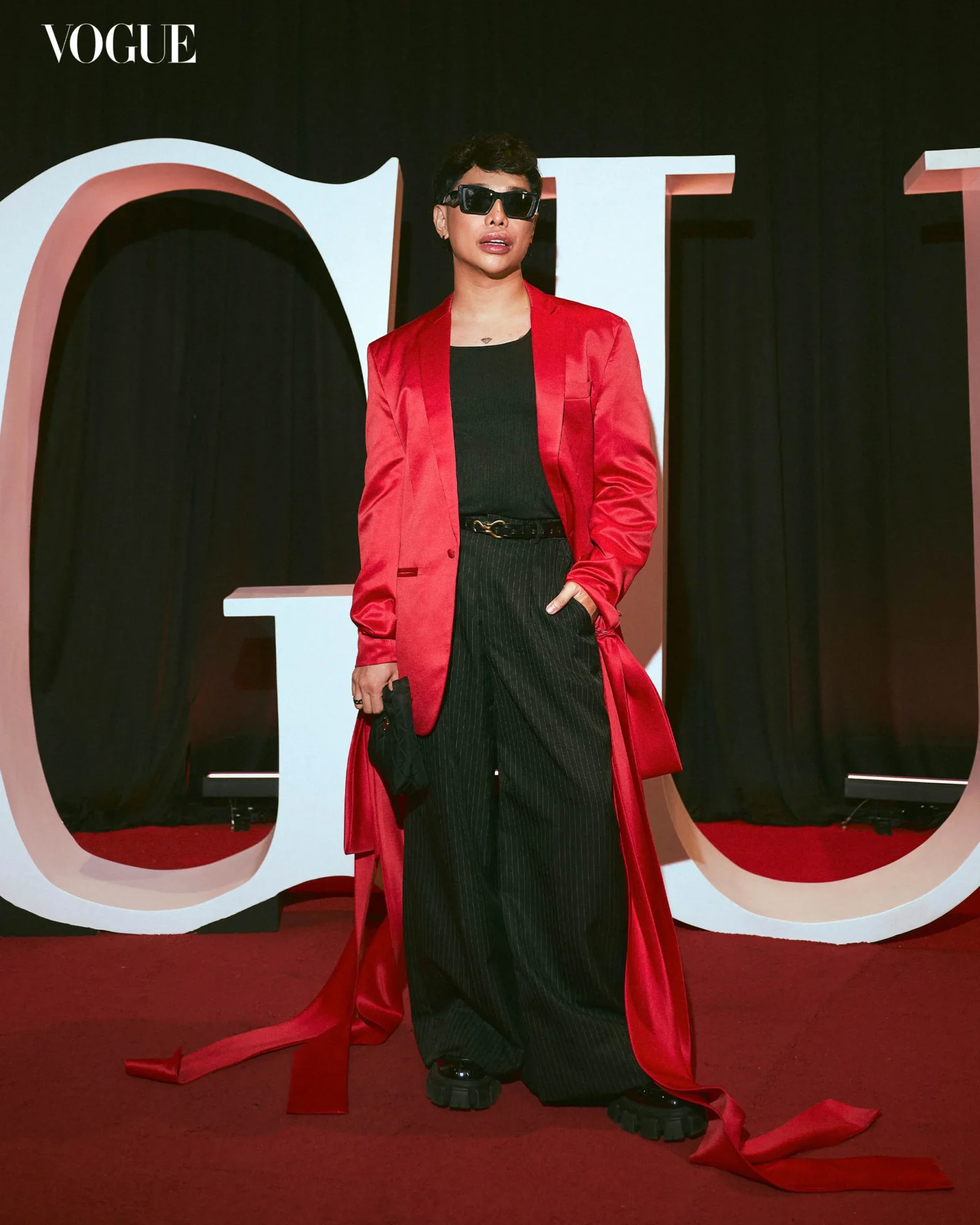 Person in a red blazer over a black top and pinstripe pants posing on a red carpet with large Vogue letters behind.