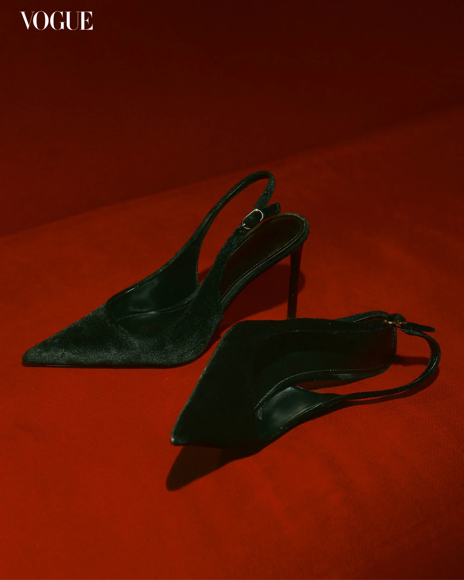Black suede high-heeled sandals with ankle straps resting on a red background, Vogue logo in the top-left corner