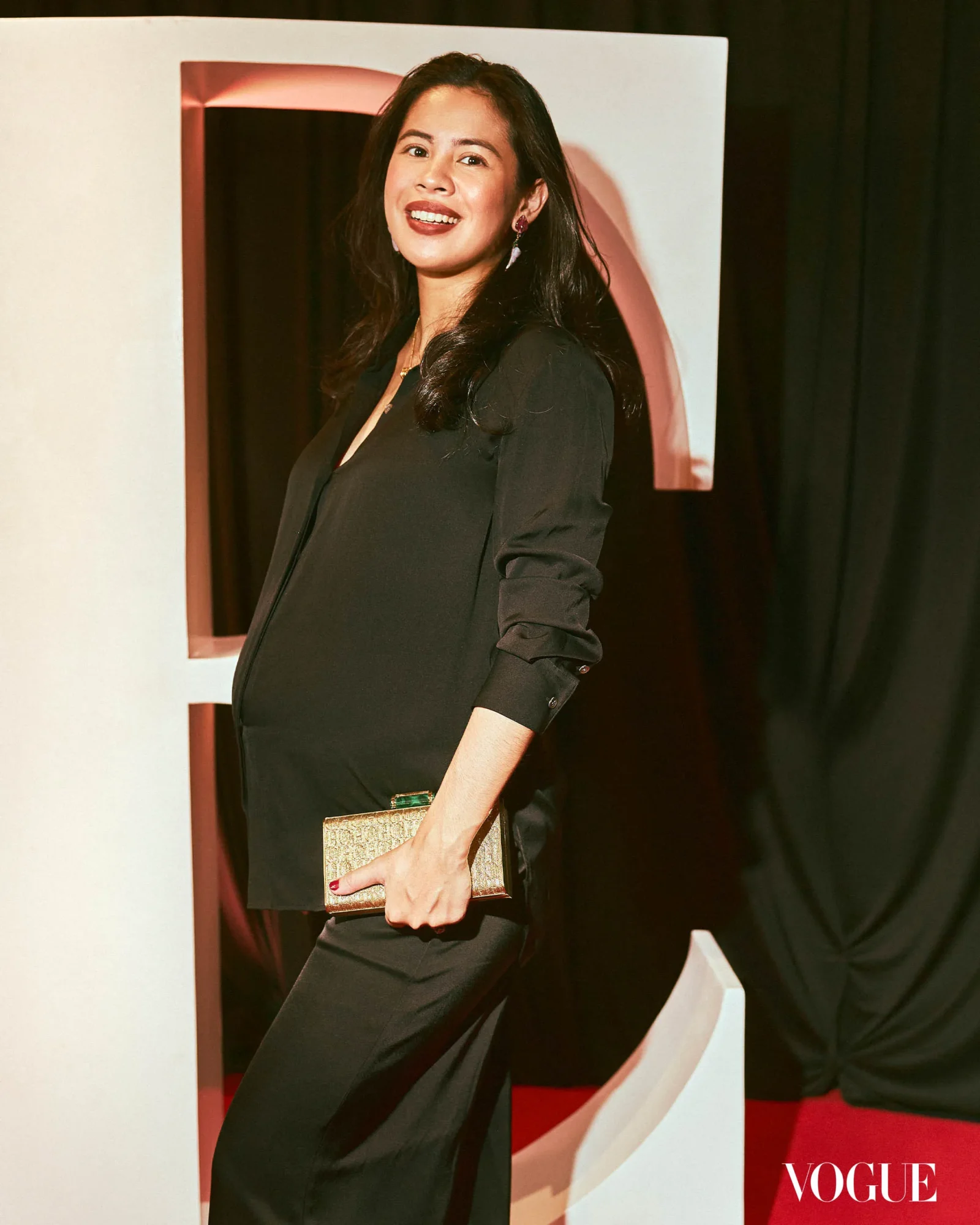 Pregnant woman in a black dress smiles while posing at a fashion event with a Vogue backdrop, holding a gold clutch.