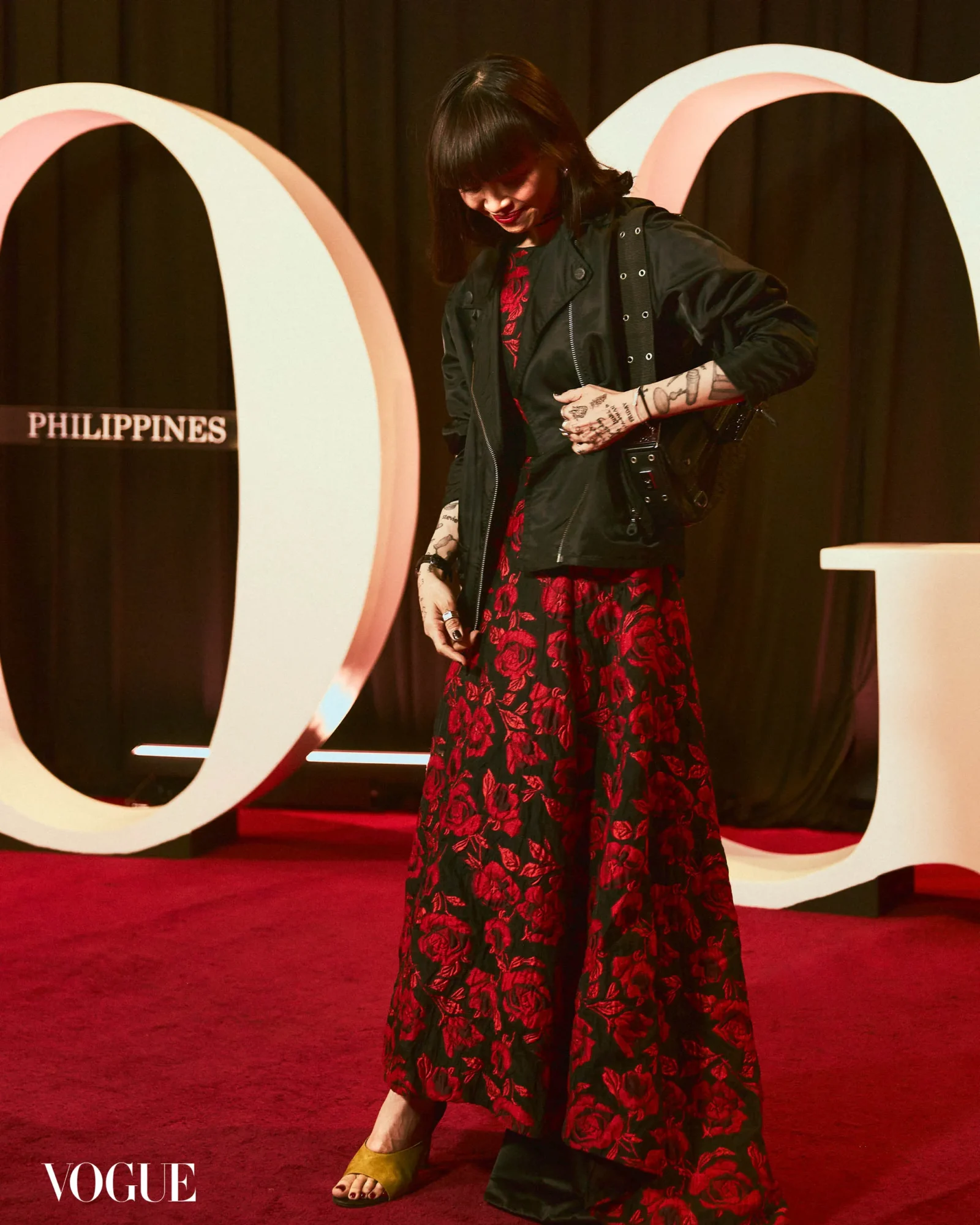 Woman with dark hair in a black jacket over a red floral dress, standing on a red carpet at a Vogue event, showing tattooed arms.