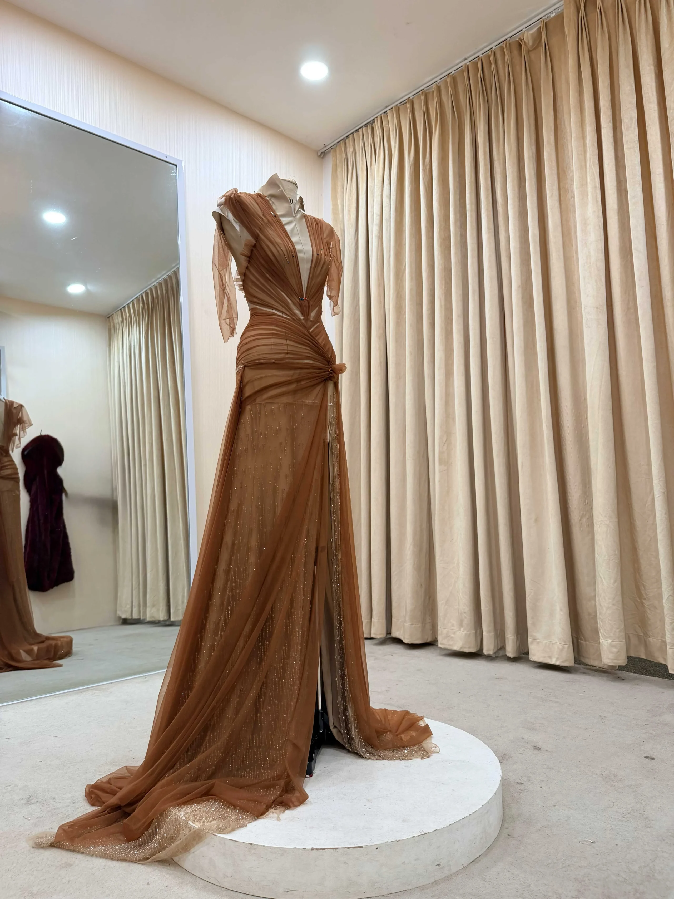Brown evening gown on a dress form, with a flowing train spilling onto a white circular platform; beige curtains in the background.