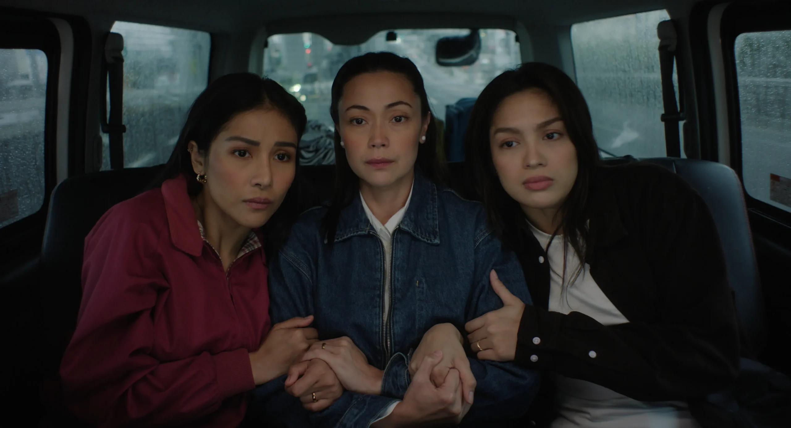 Three women sit closely in the backseat of a car, holding hands and looking toward the camera with serious expressions.