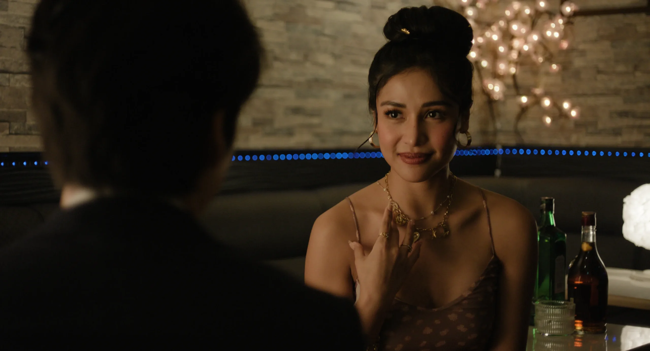 Woman in a brown dress with gold jewelry sits at a dimly lit bar, facing a man whose silhouette is in the foreground.