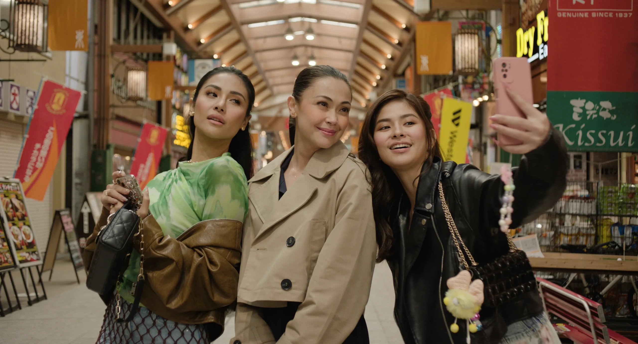 Three women posing for a selfie in a busy indoor arcade with colorful banners and shops visible in the background, smiling at the camera.