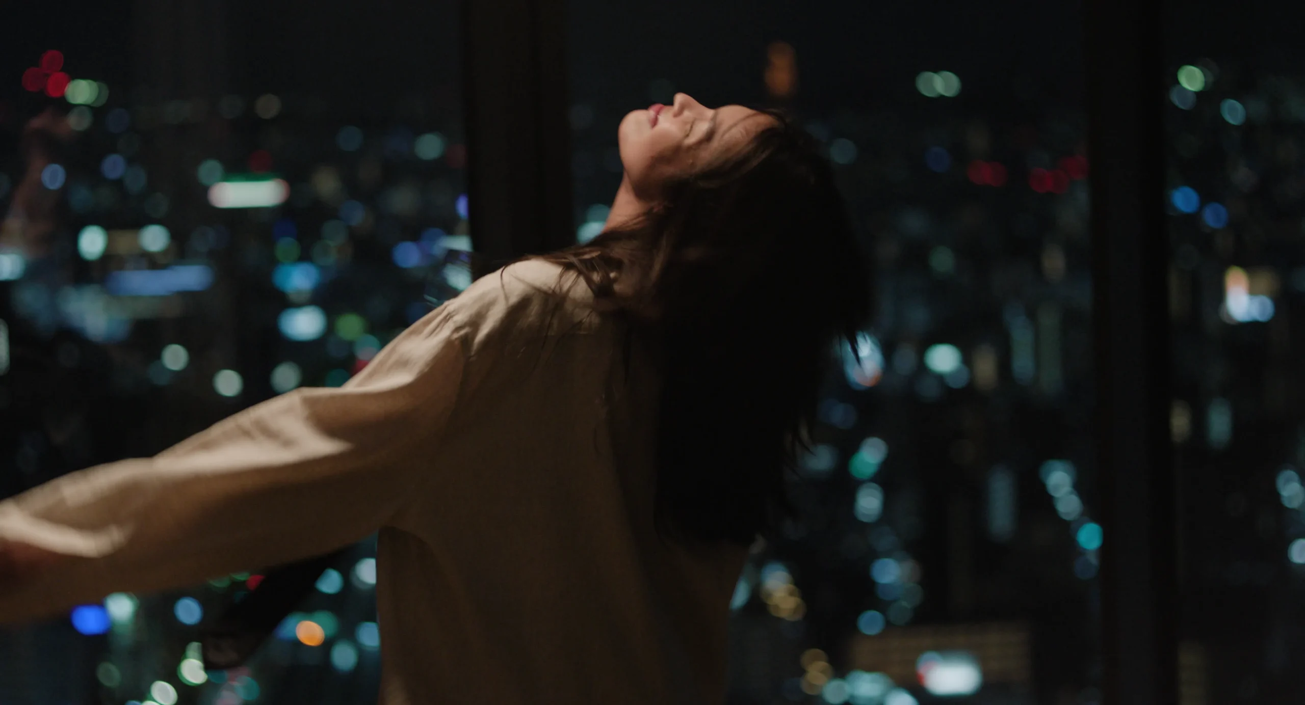 Person with arms outstretched at night, overlooking a cityscape with blurred colorful lights behind.