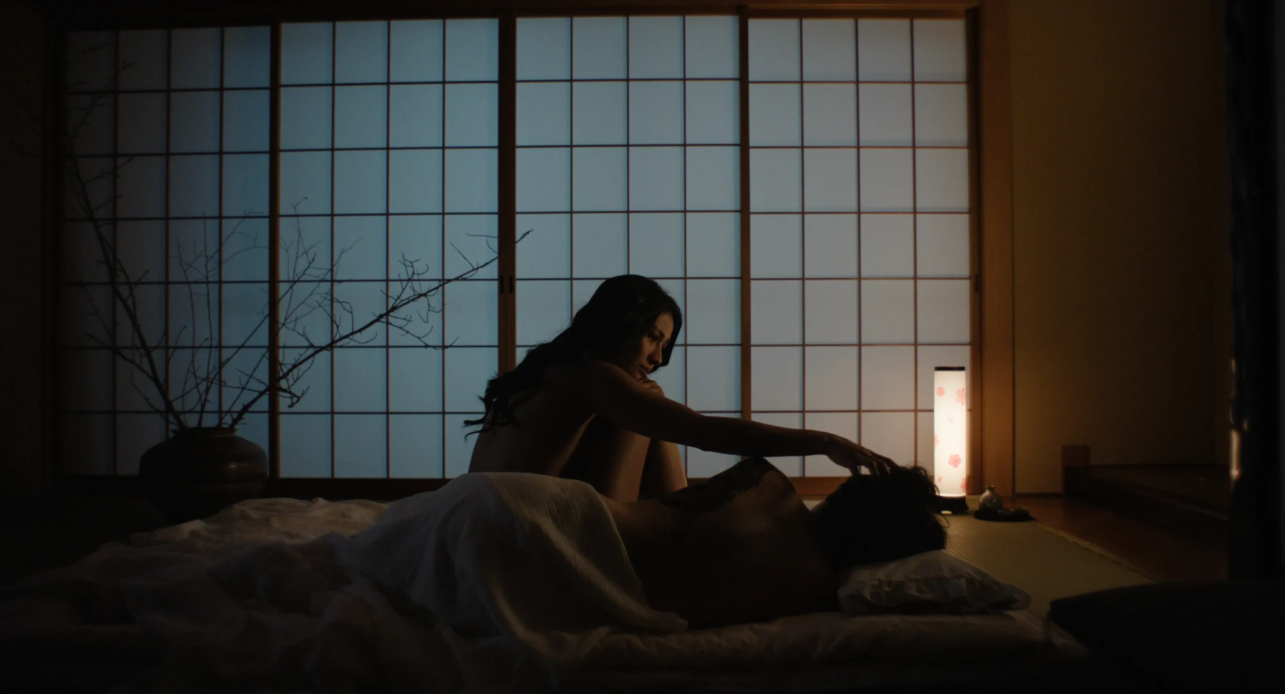 Dim bedroom with a couple on a bed by a large grid window; a small lamp glows on the right.