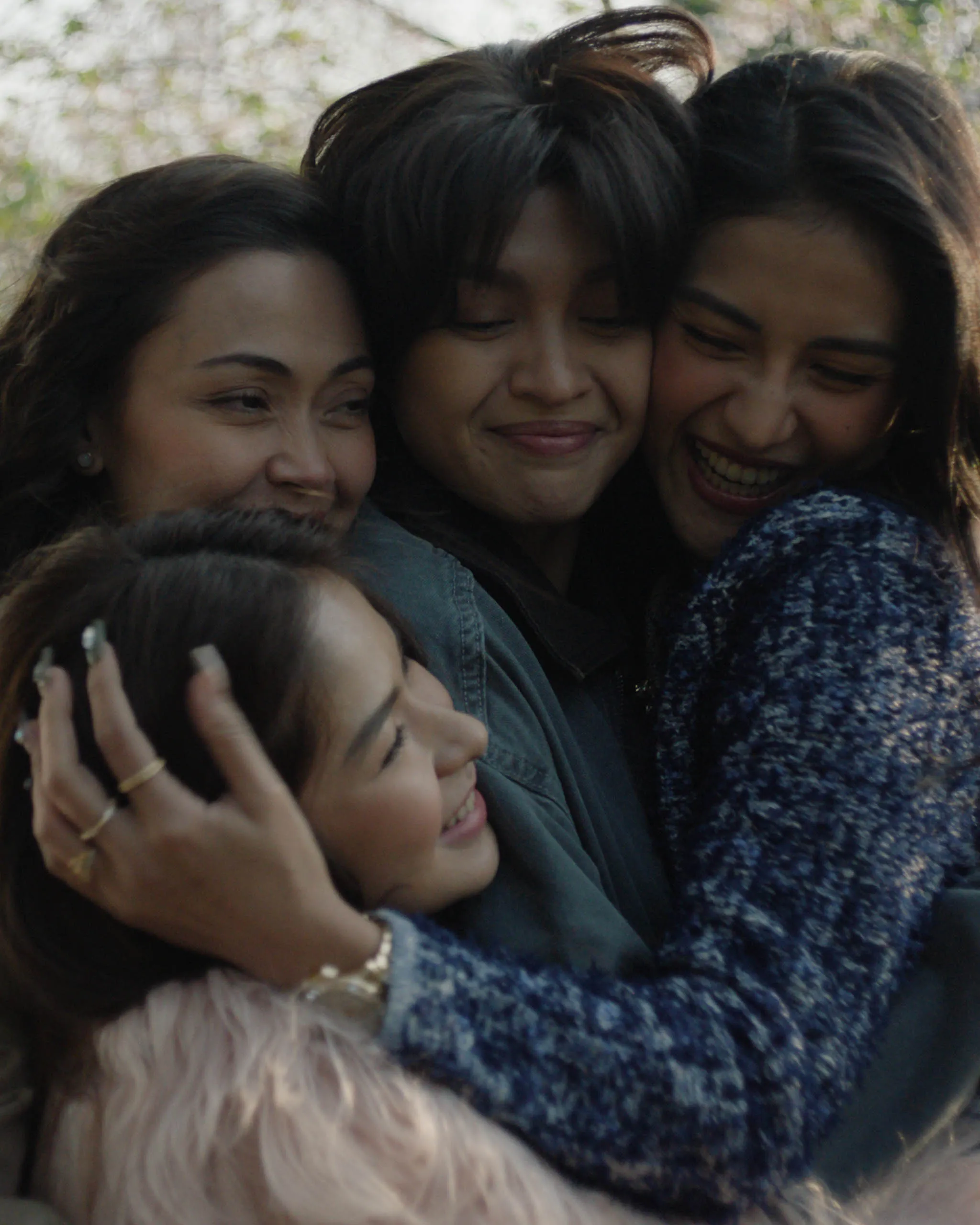Three women hugging and smiling outdoors, close crops of their faces.