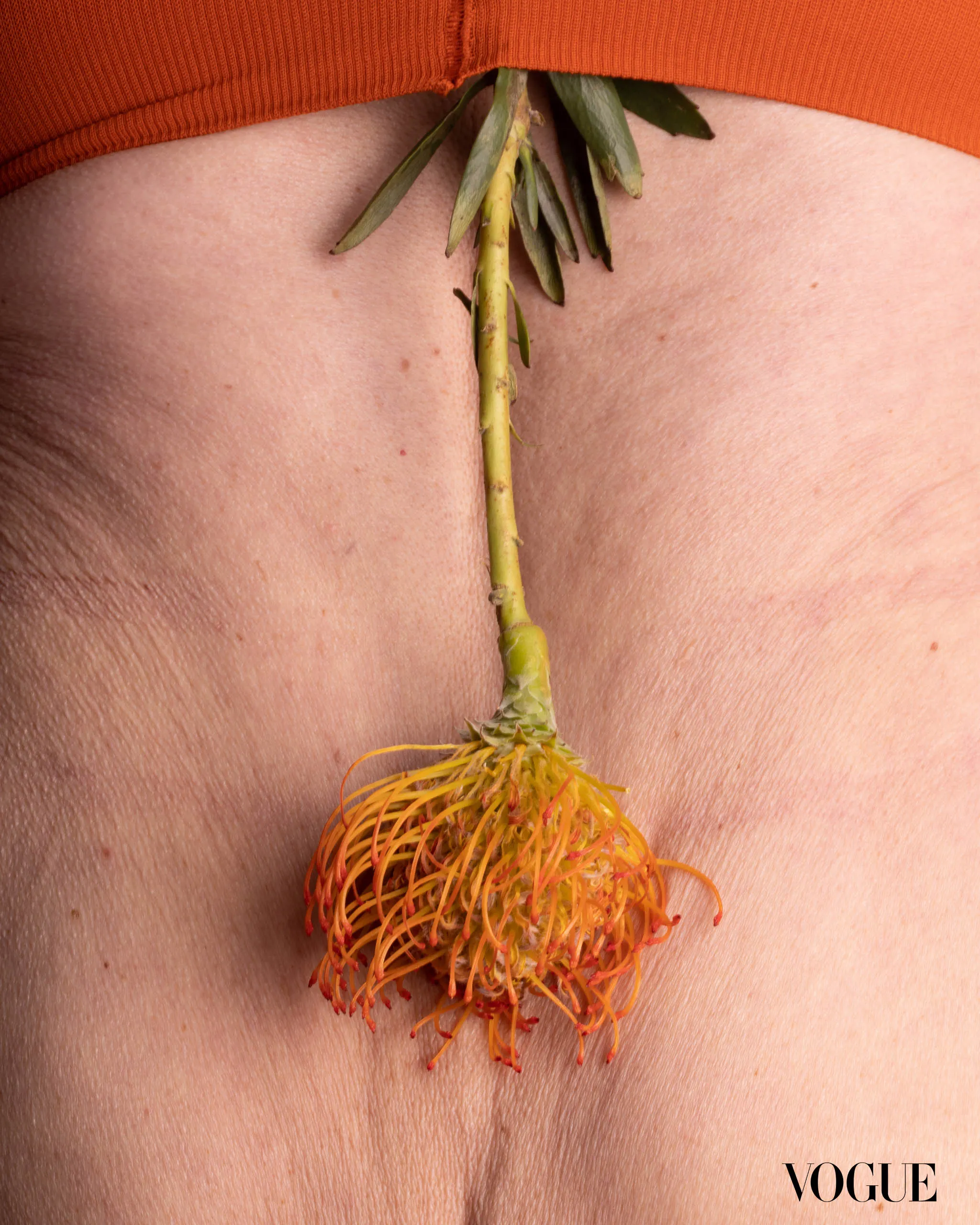 Close-up of a fashion image showing a yellow-orange floral pendant hanging between a model's cleavage with an orange top and Vogue watermark in the corner.