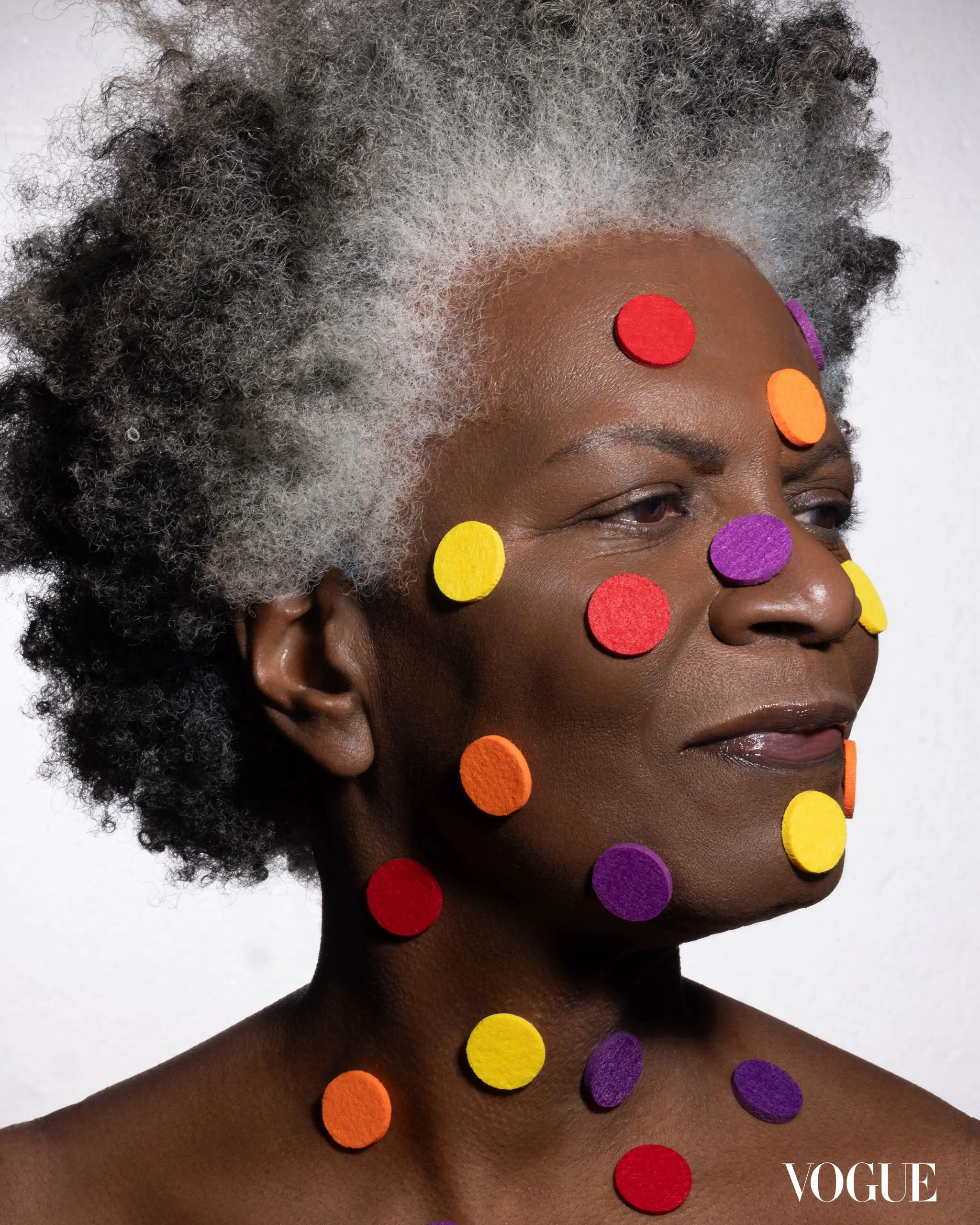 Close-up portrait of a dark-skinned person with bright circular stickers on the face and neck, Vogue logo visible in the corner.