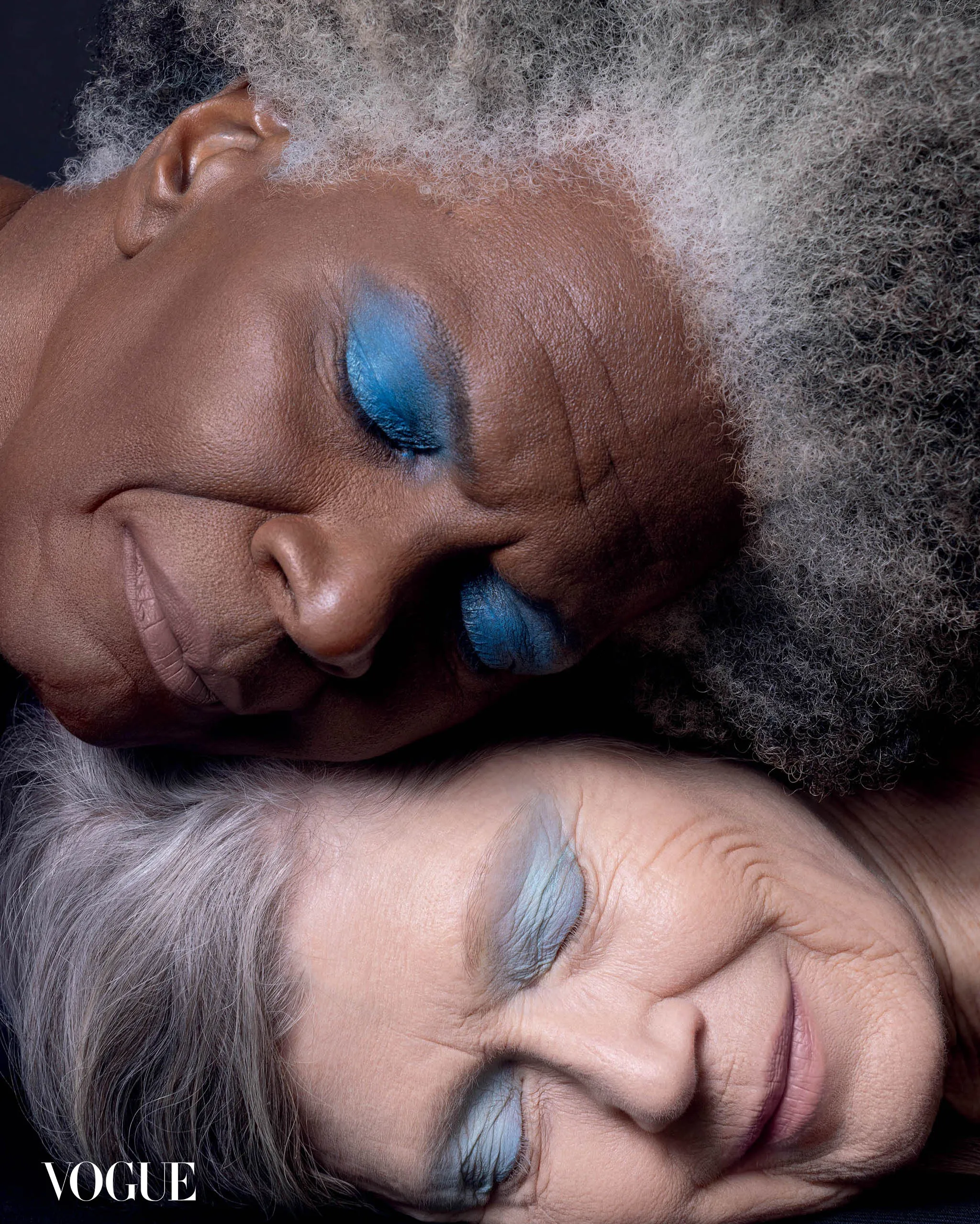 Close-up portrait of two elderly women resting their heads together, one with vivid blue eye makeup; Vogue logo visible.