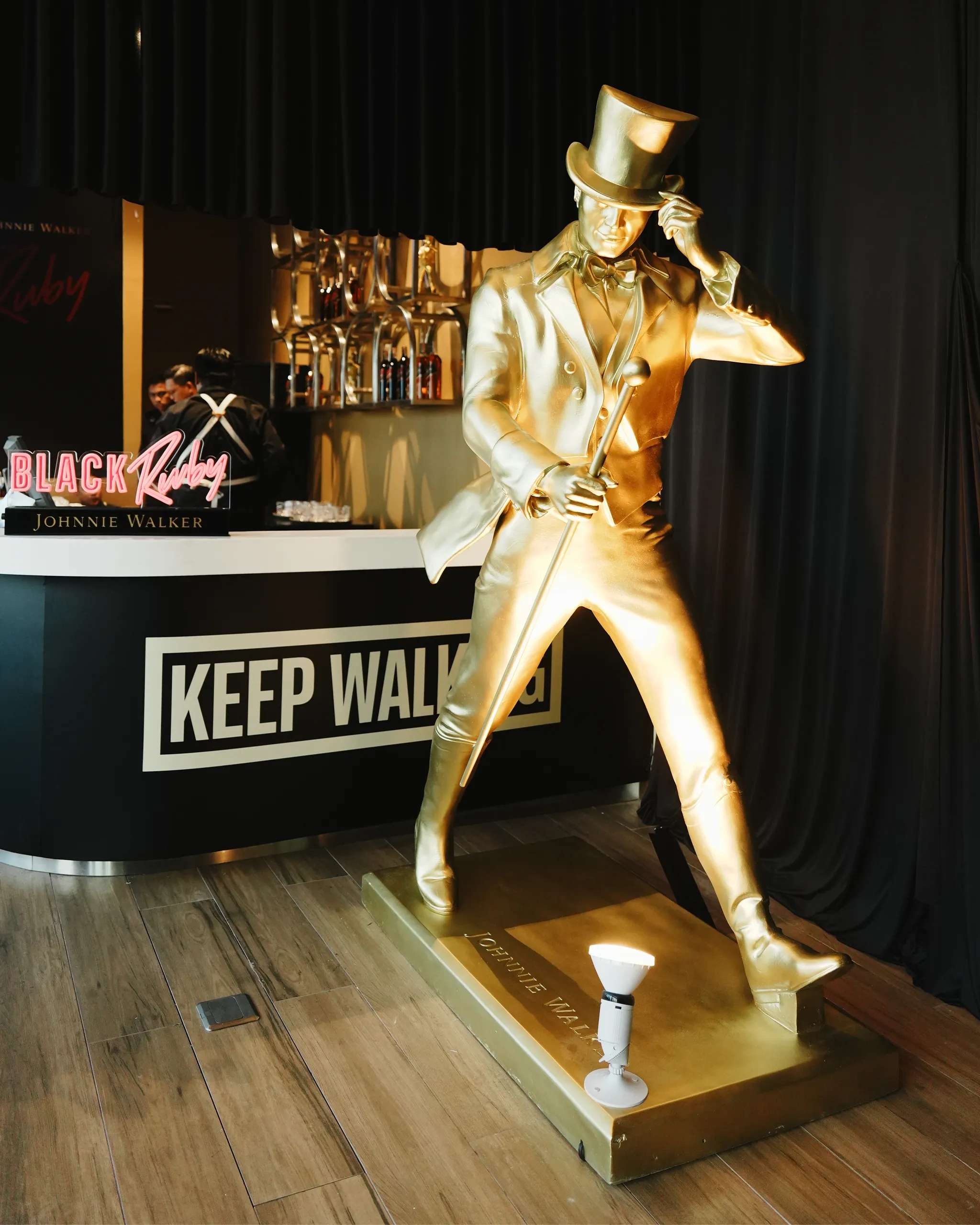 Golden statue of a man in a suit and top hat with a cane, part of a Johnny Walker Keep Walking display in a bar setup.