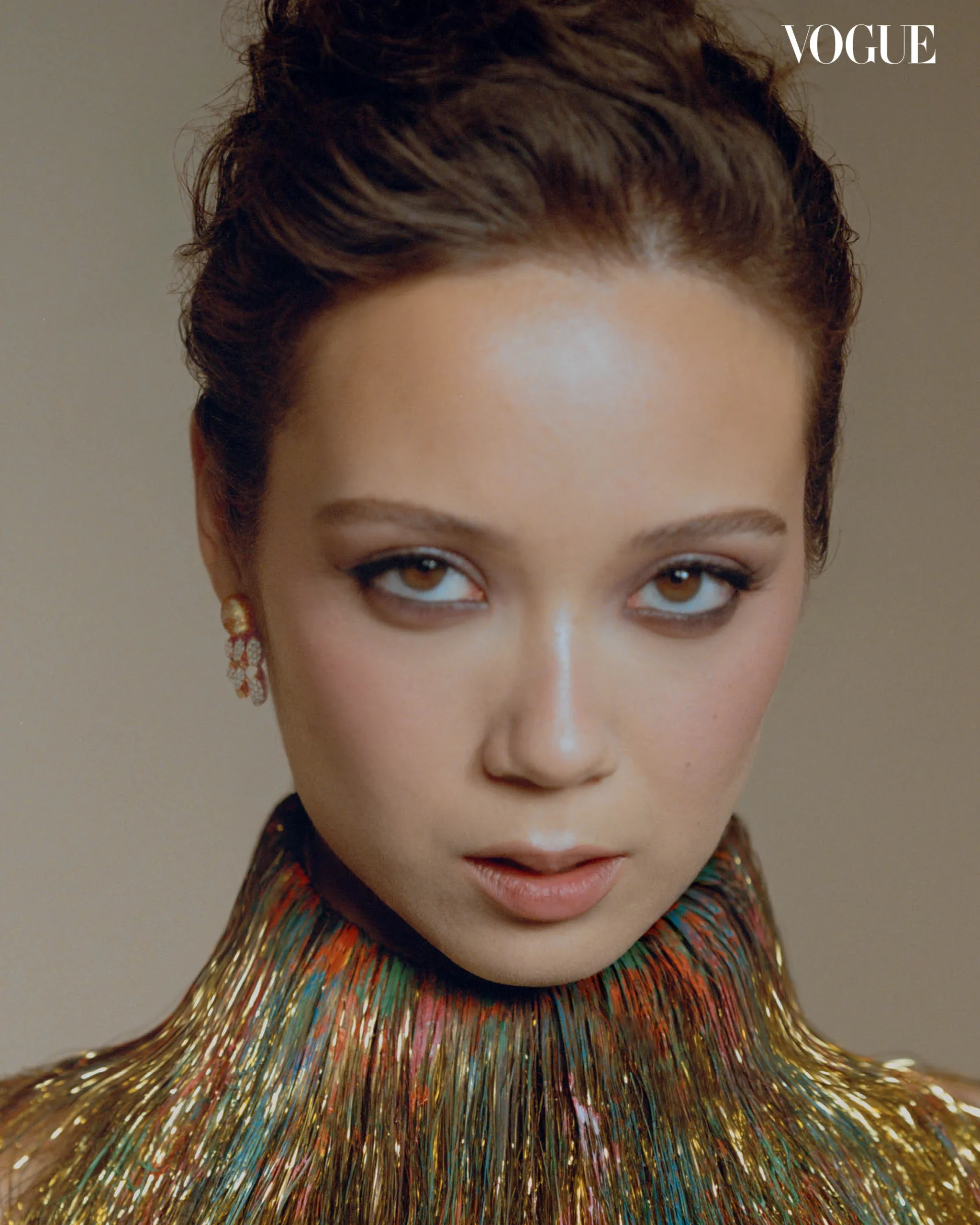 Close-up of a woman with brown eyes and glossy lips wearing a multicolored metallic fringe necklace; Vogue logo in the corner.