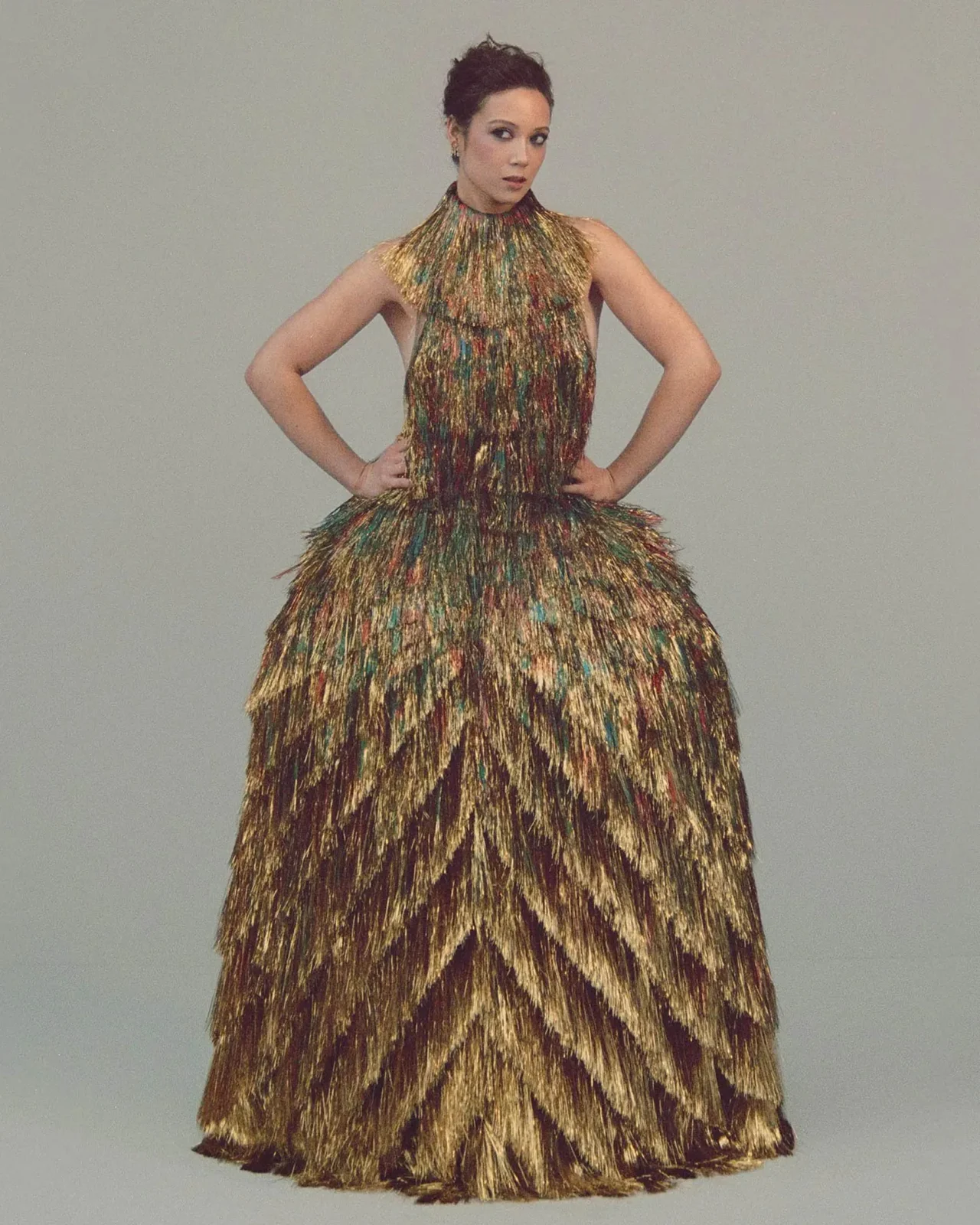 Model posing in a gold, tiered, feather-like gown with hands on hips against a muted gray backdrop.