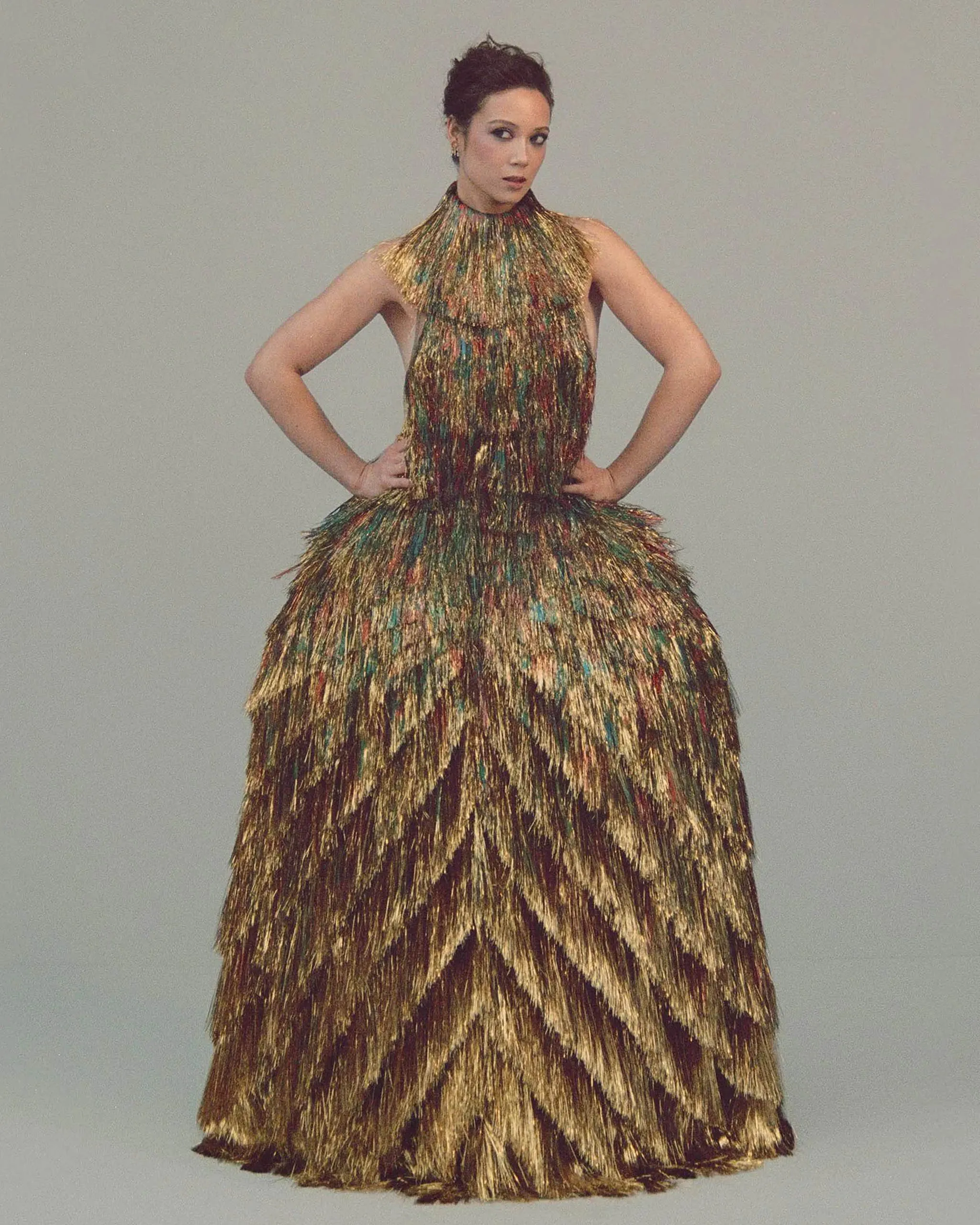 Model wearing a shimmering gold multicolored fringe gown with hands on hips against a plain background.