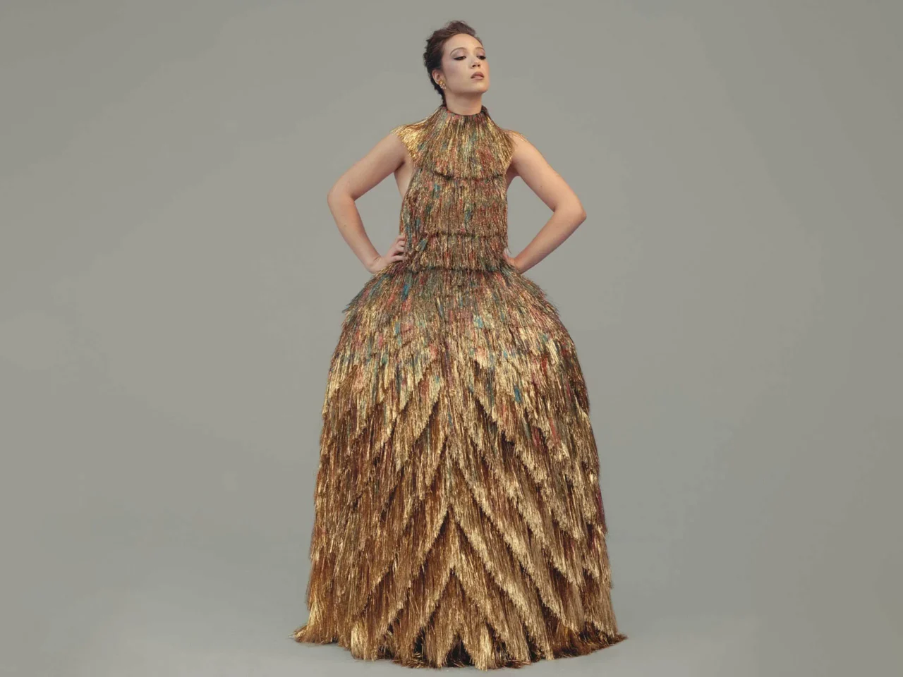 Model posing in a gold, tiered, feather-like gown with hands on hips against a muted gray backdrop.