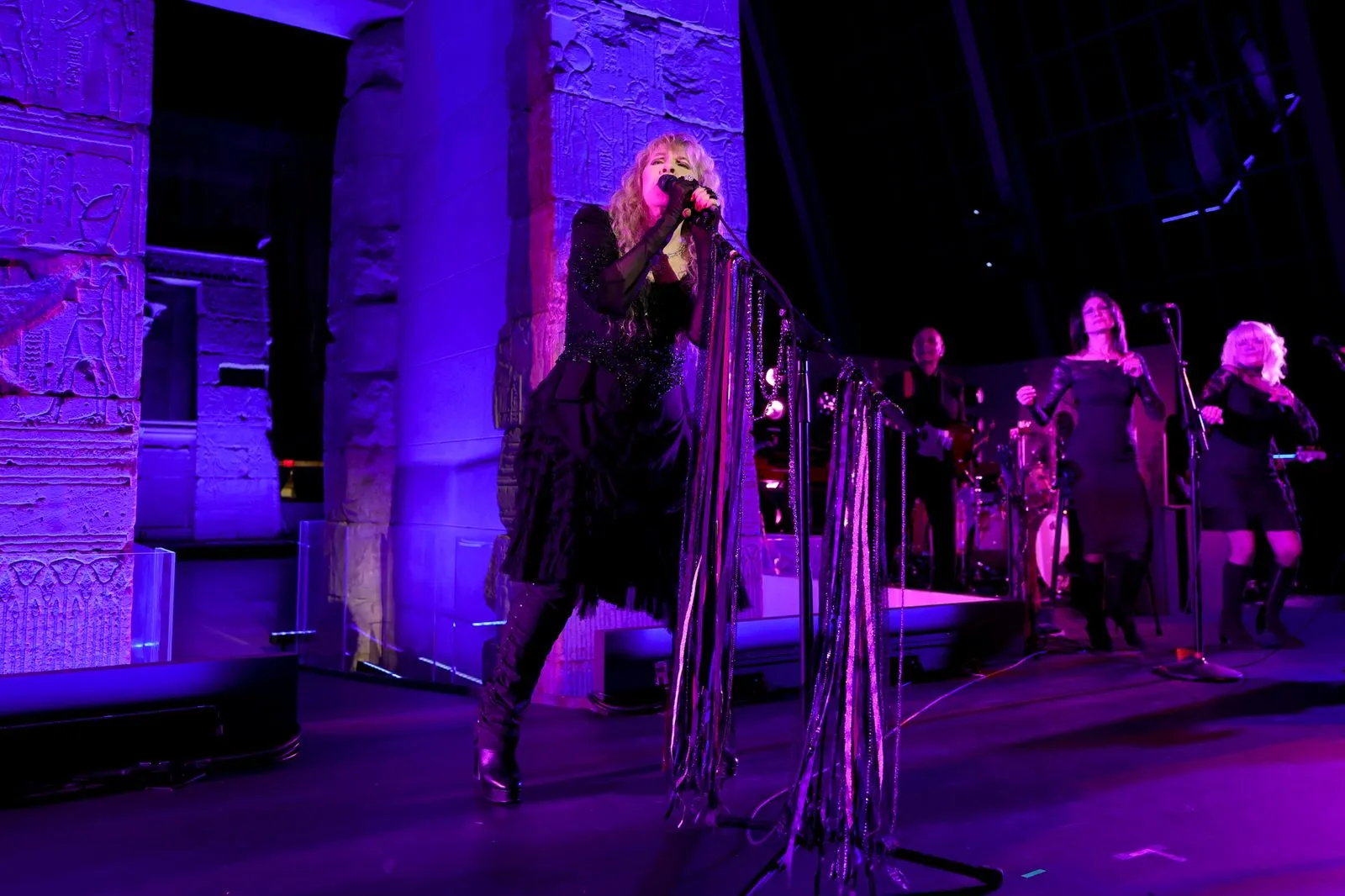 Female lead singer in black outfit sings into a microphone on a purple-lit stage, fringed streamers hanging from the mic stand, with a band in the background.