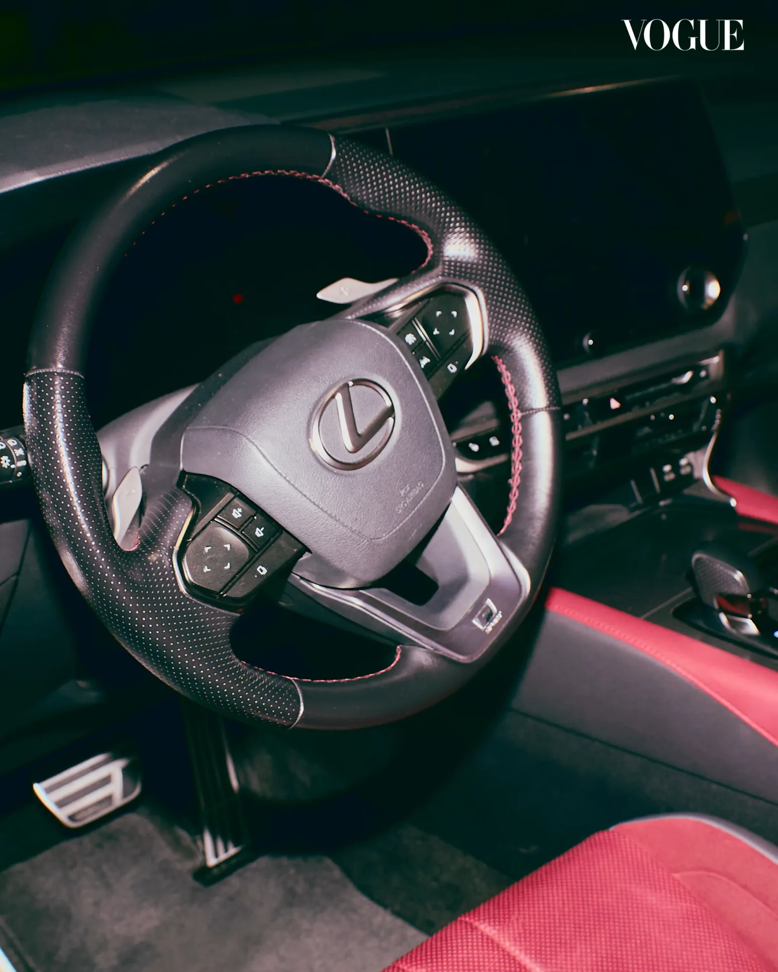 Steering wheel with textured grip and control buttons inside a dark car cabin, Vogue logo in the top-right corner