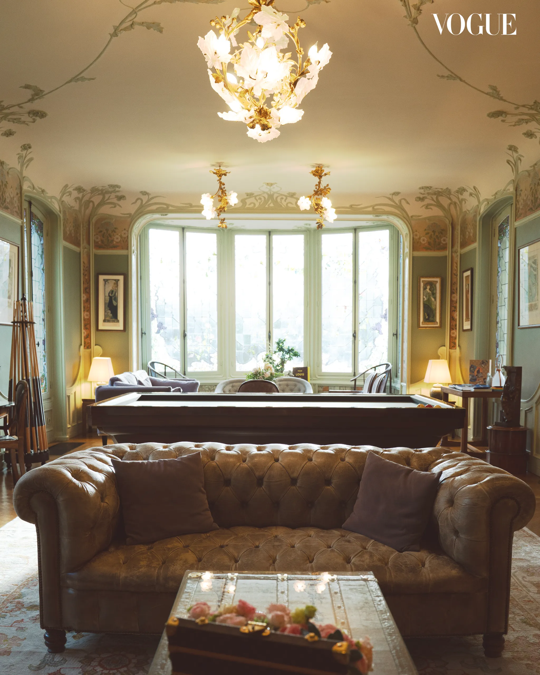 Elegant living room with a tufted brown sofa facing a glass coffee table; soft light from a large bay window and a decorative ceiling chandelier (Vogue watermark).