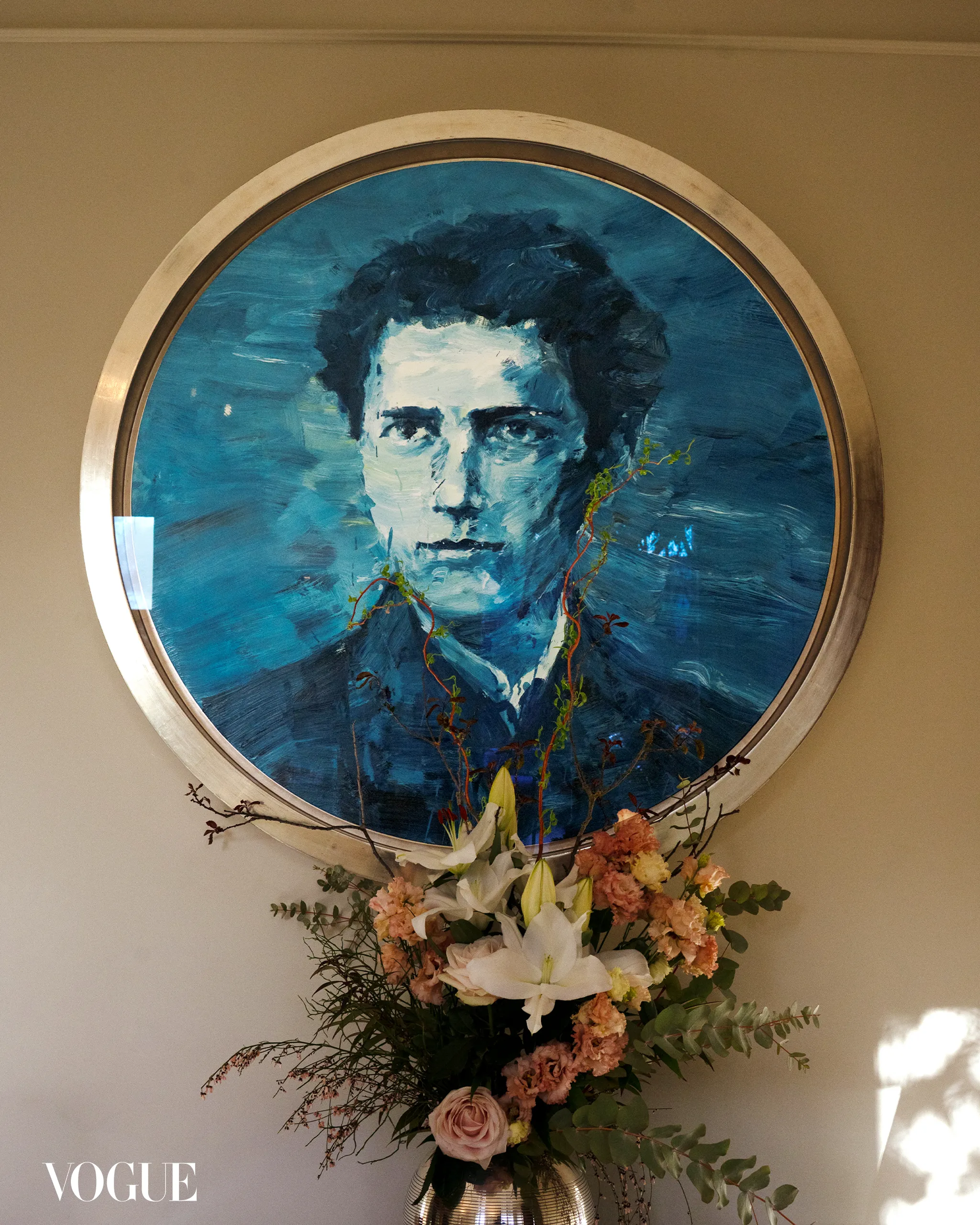 Circular blue portrait painting in a metal frame mounted on a beige wall, with a floral arrangement of lilies and pink blooms beneath. Vogue logo visible in lower left corner.