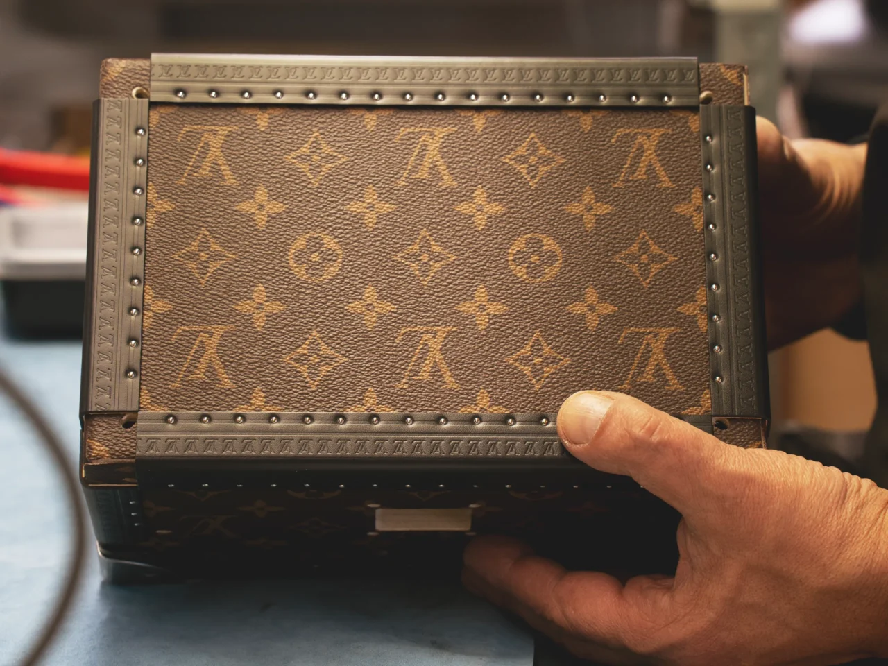 Close-up of a brown Louis Vuitton monogram bag being held along the bottom edge by a hand, showing riveted trim.