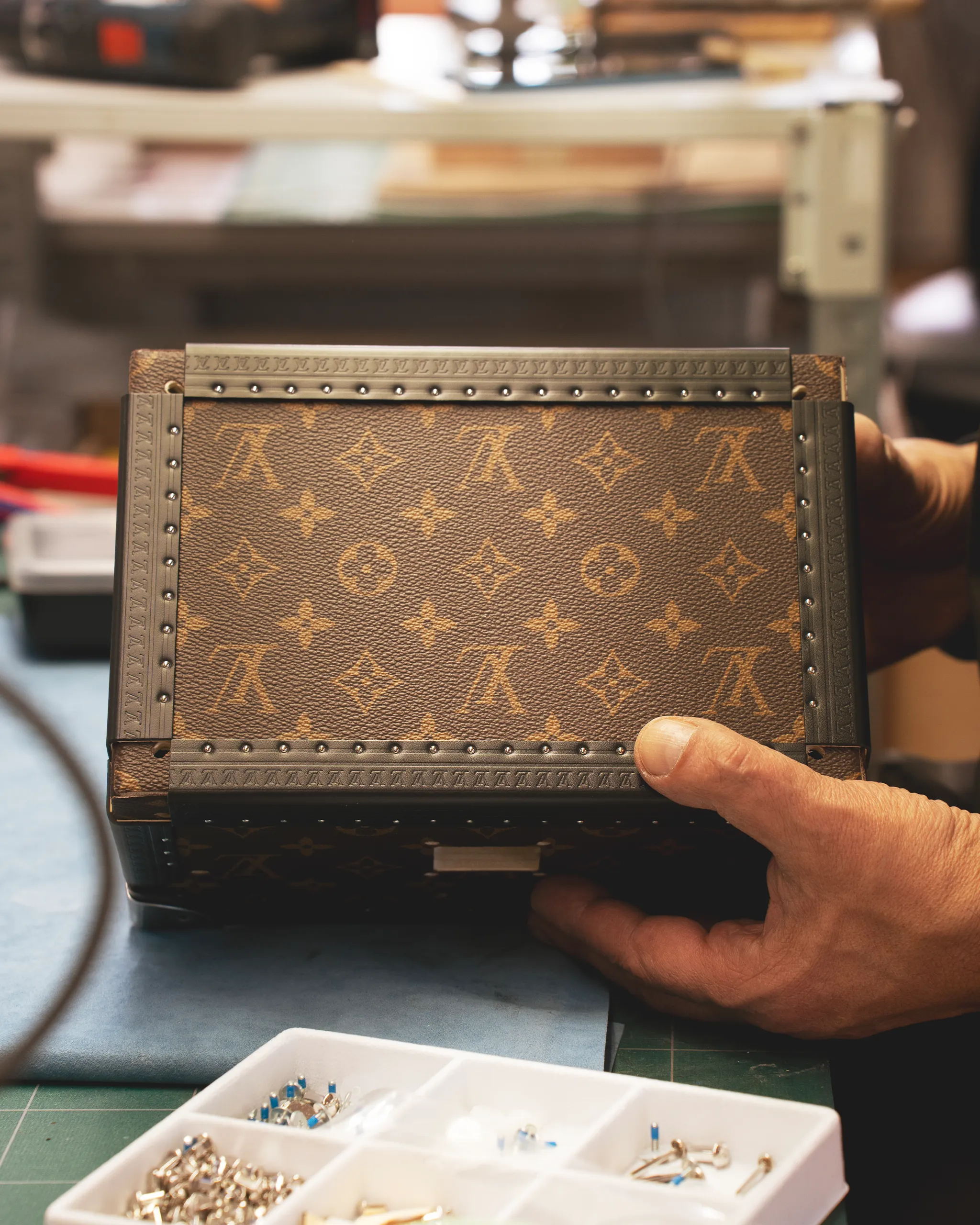 Hands hold a brown Louis Vuitton monogram trunk with metal rivets on a workbench, next to a tray of small jewelry pieces.