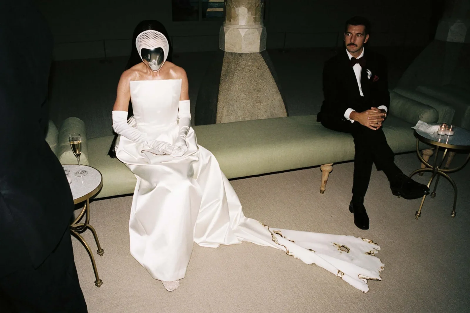 Person in a white strapless wedding dress and a futuristic helmet sits on a pale sofa, with a man in a black tuxedo beside them in a dim room; a champagne glass rests on a small table nearby.