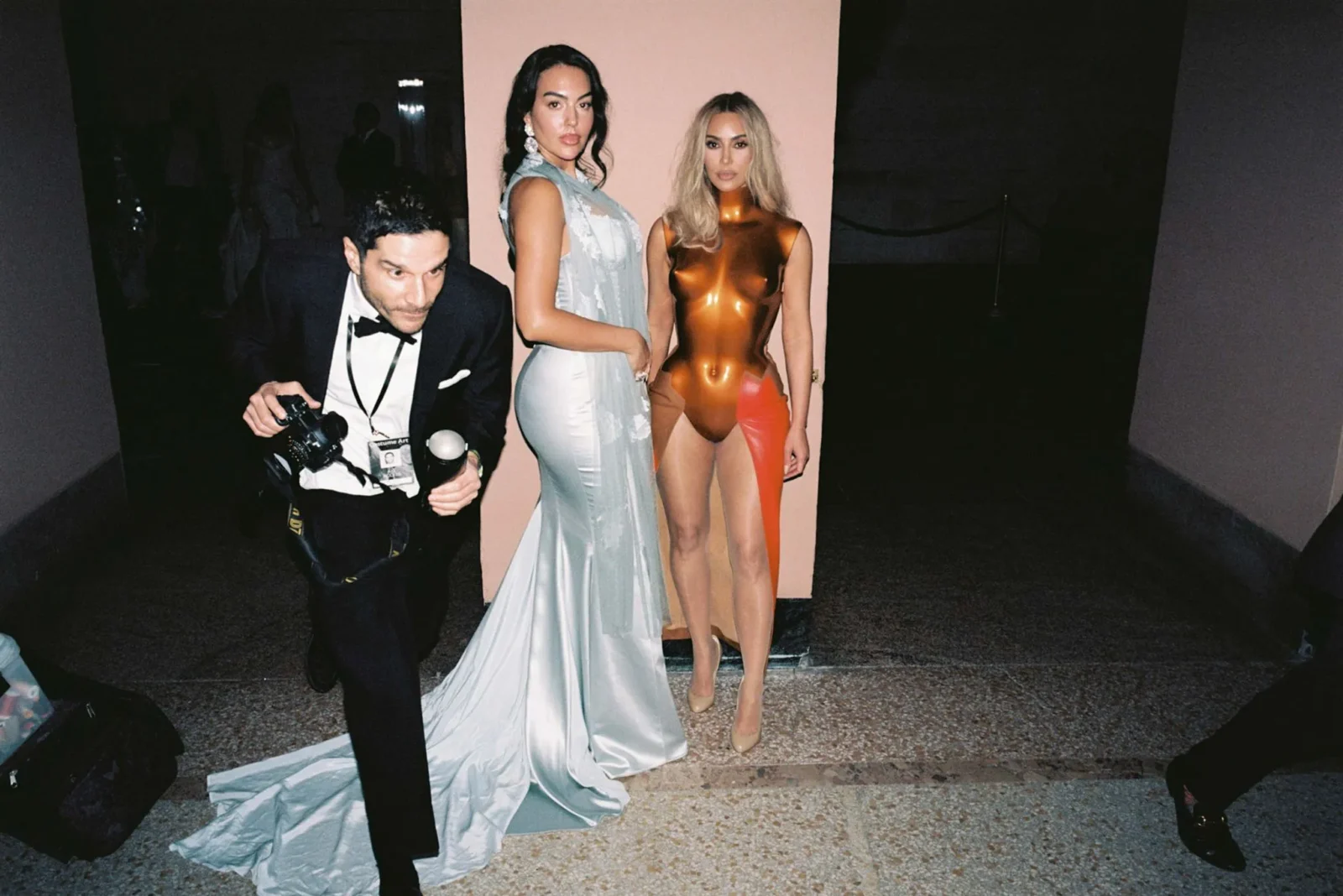 Photographer in a tuxedo crouches with a camera while two models pose in front of a pale pink backdrop, one in a sky-blue gown and the other in a copper metallic bodysuit.