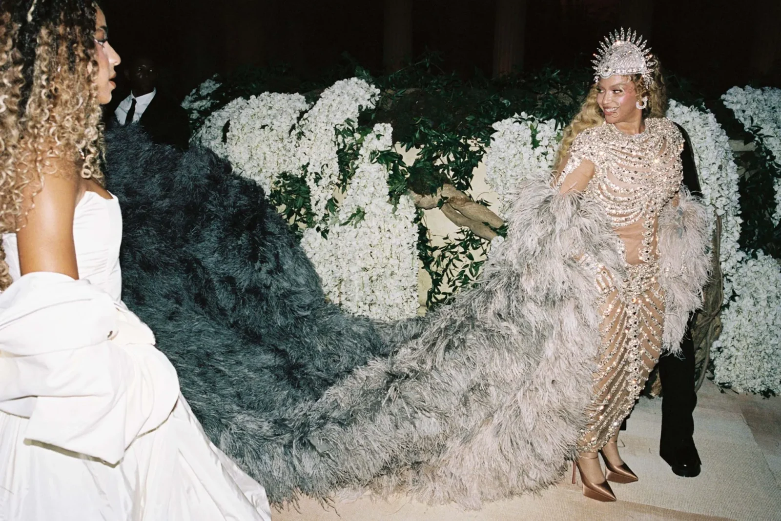 Glamorous woman in a pearl-embellished gown and crown with a feathered train stands beside a guest in a white dress among white flower arrangements.