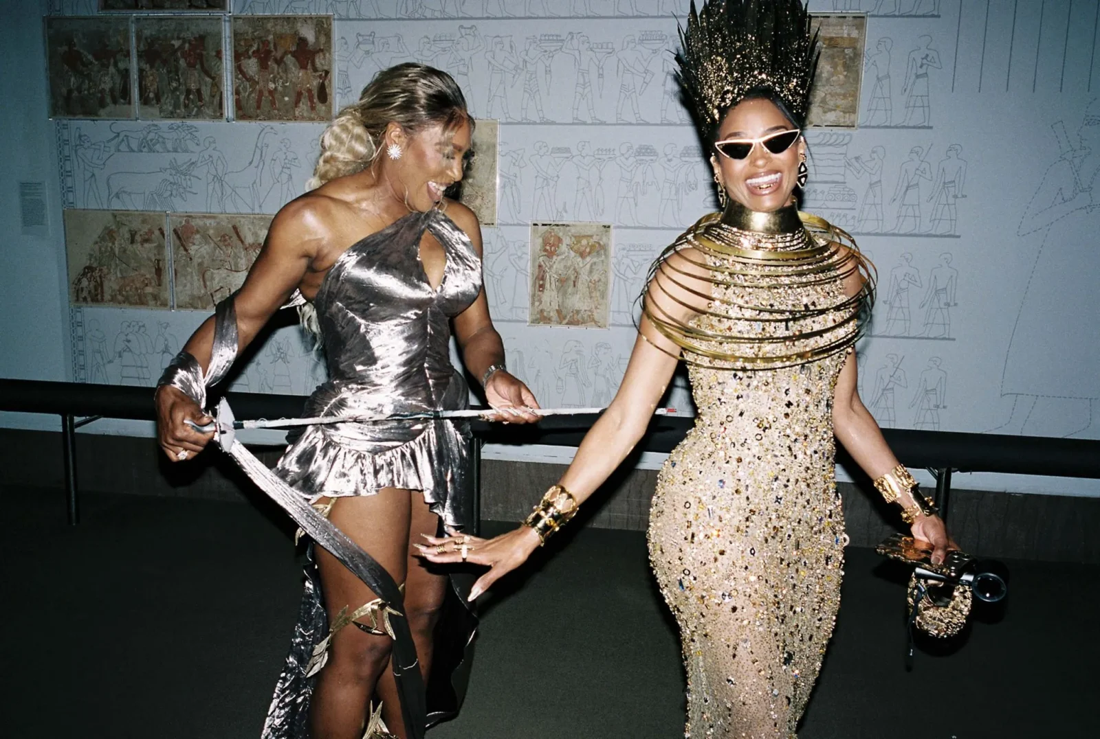 Two women in metallic gowns dance together, smiling and pulling a ribbon at a party with a mural backdrop behind them