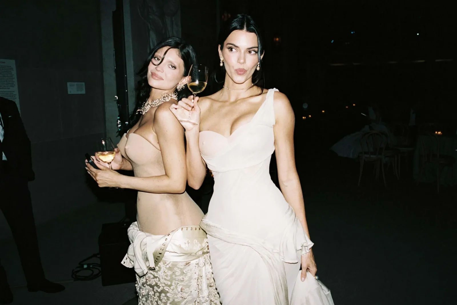 Two women in elegant evening dresses pose with wine glasses at a nighttime party, smiling for the camera.