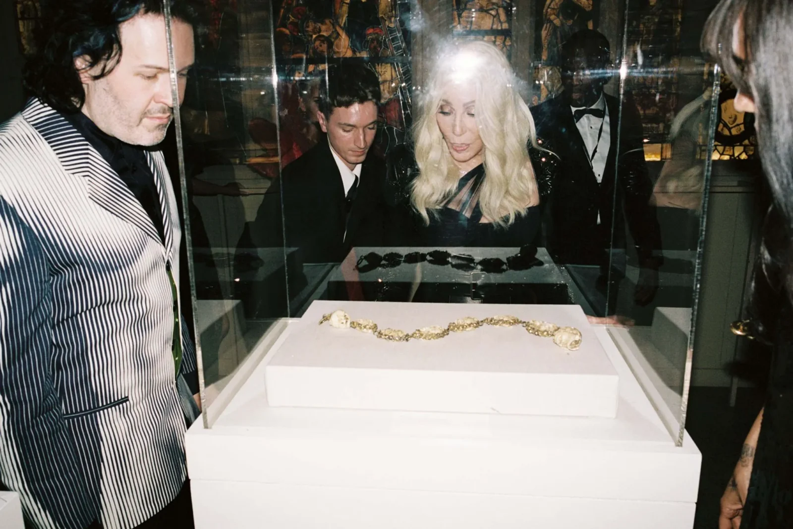 People in a museum-like setting gaze at a glass display case containing gold jewelry laid out on a white tray inside a case.