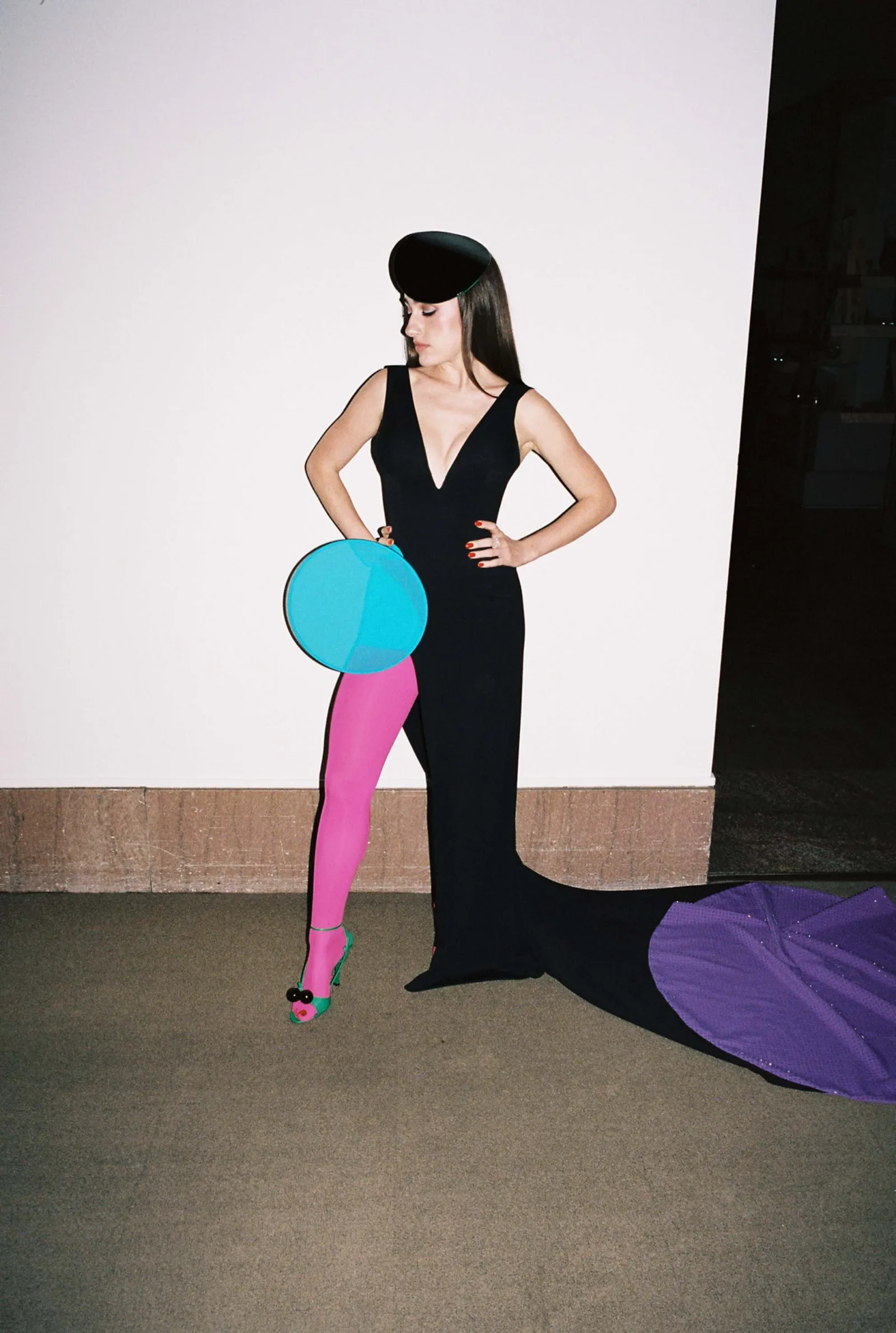 Fashion model in a black sleeveless gown with a deep V-neck, pink tights, green heels, and a black hat, posing with a bright blue circular prop against a white wall.