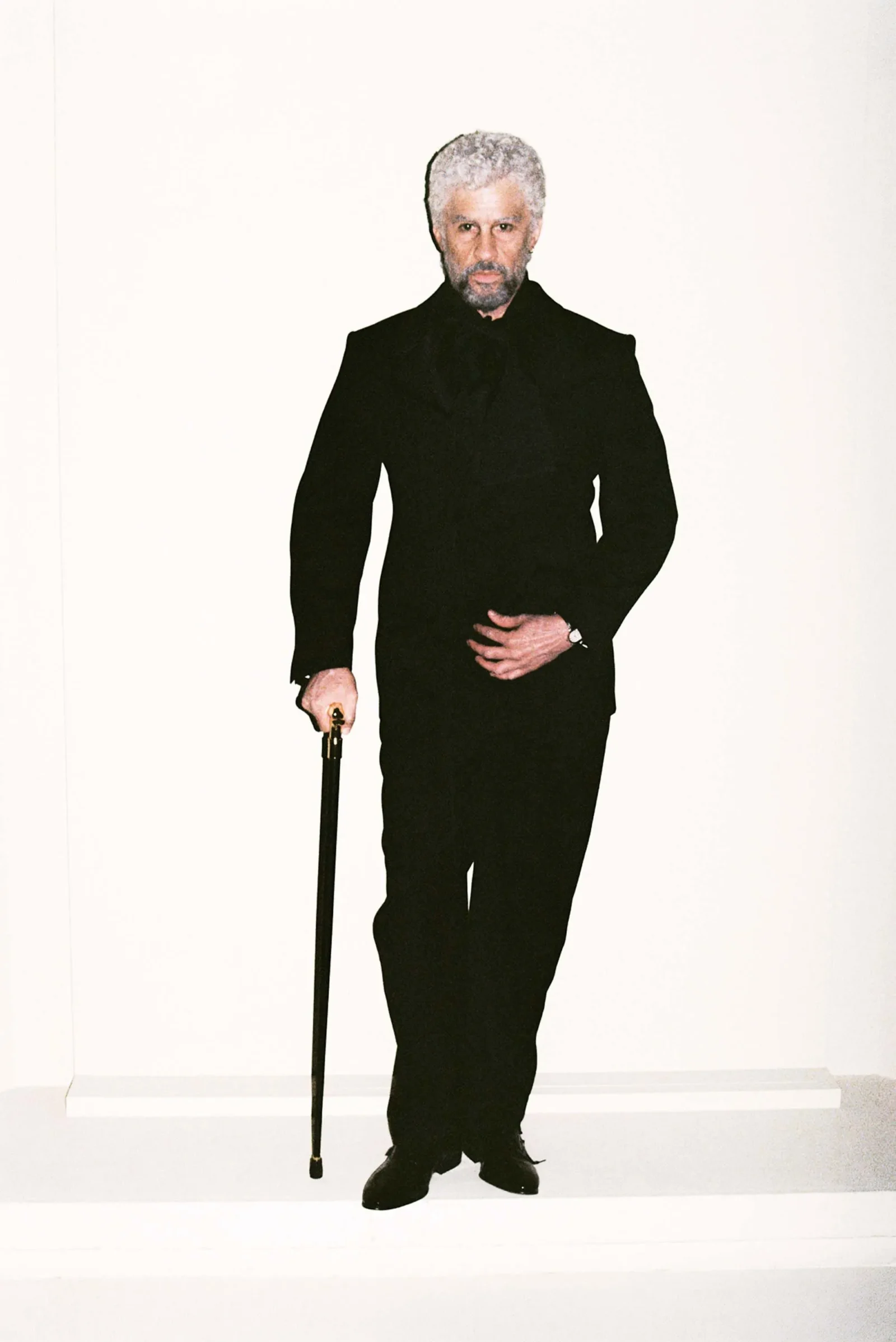 Older man with gray hair and beard, wearing a black suit, standing against a white background and holding a cane.