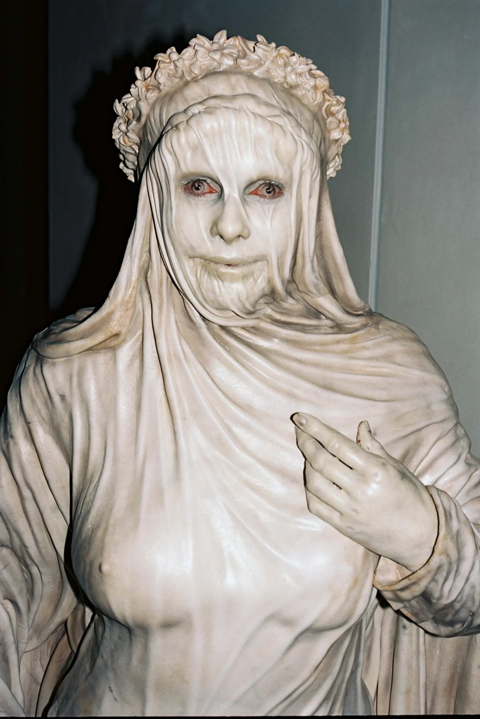 White marble statue of a veiled woman wearing a flower crown, with a calm, pale face and red-tinted eyes, her hand raised in a gesture.