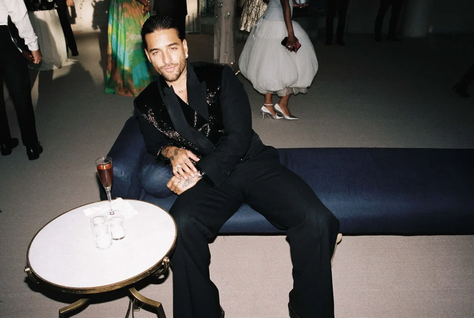 Man in a black sequined blazer sits on a blue couch at a formal party, with a round table and drinks in the foreground.