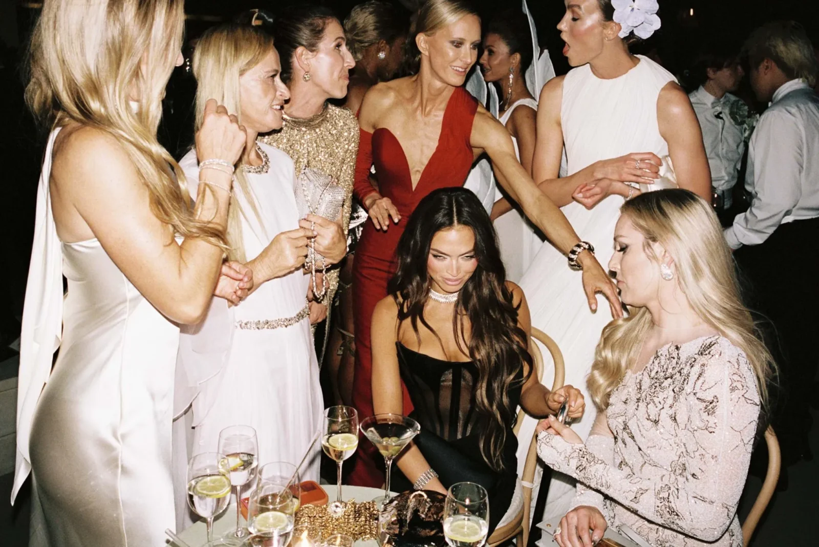 Group of women in formal dresses gathered around a table with cocktails at a party, smiling and chatting at a gala event.