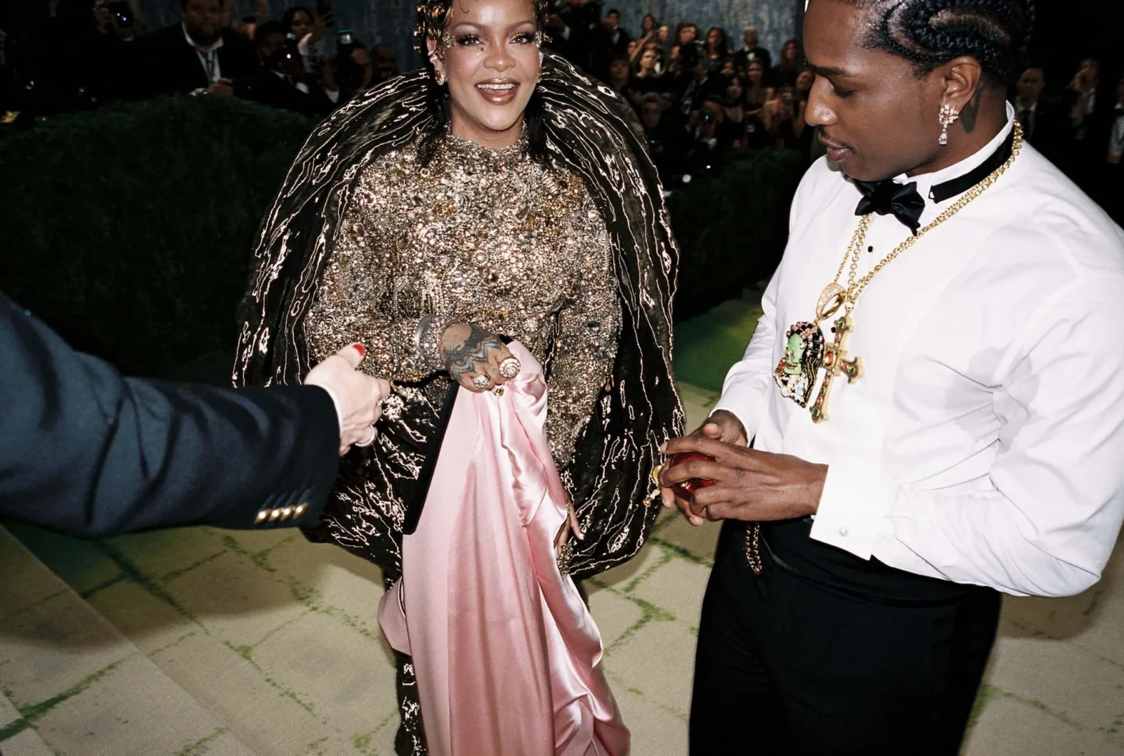 Woman in a sparkling gold beaded gown and cape shakes hands with a man in a white shirt and gold chain at a formal event; photographers in the background.