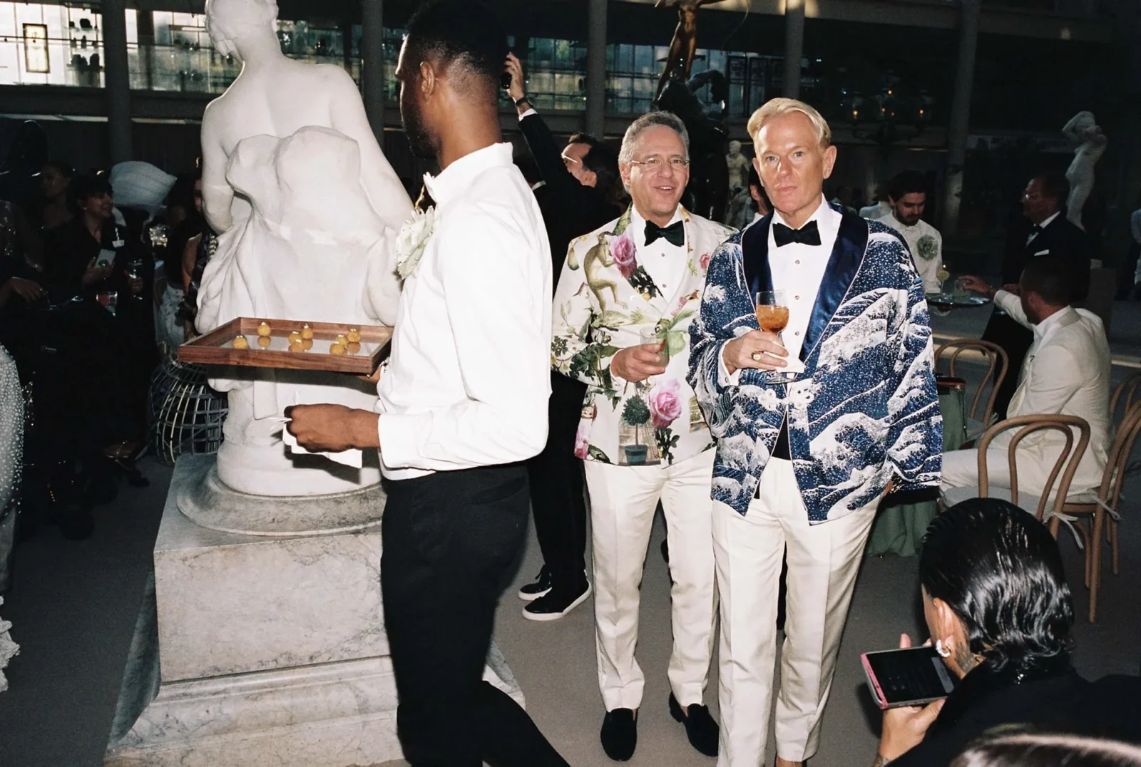 Two men in bold tuxedos posing with drinks at a formal party, statues and guests in the background.