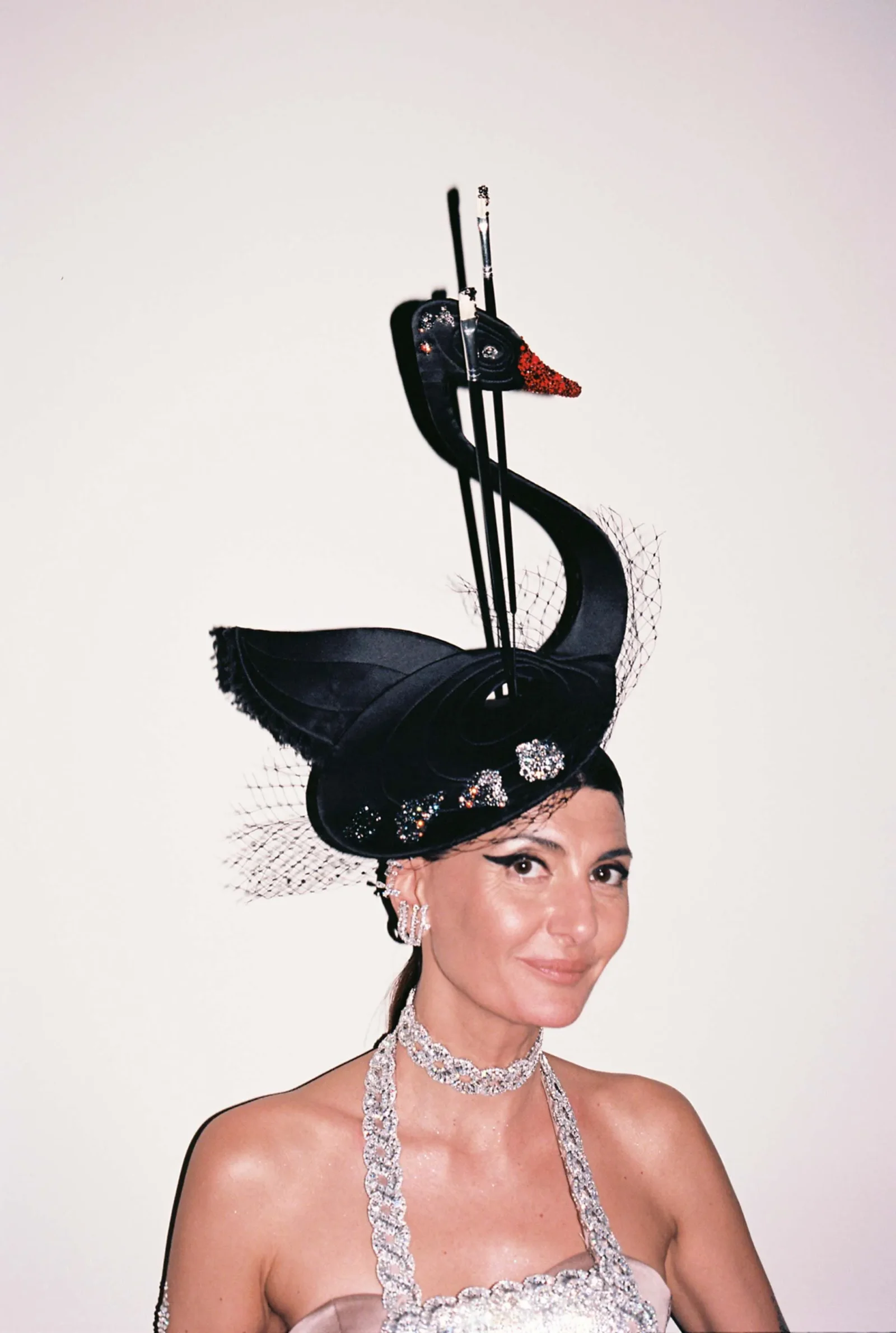 Portrait of a woman wearing a dramatic black sculptural hat with a red beak, fishnet veil, and diamond jewelry.