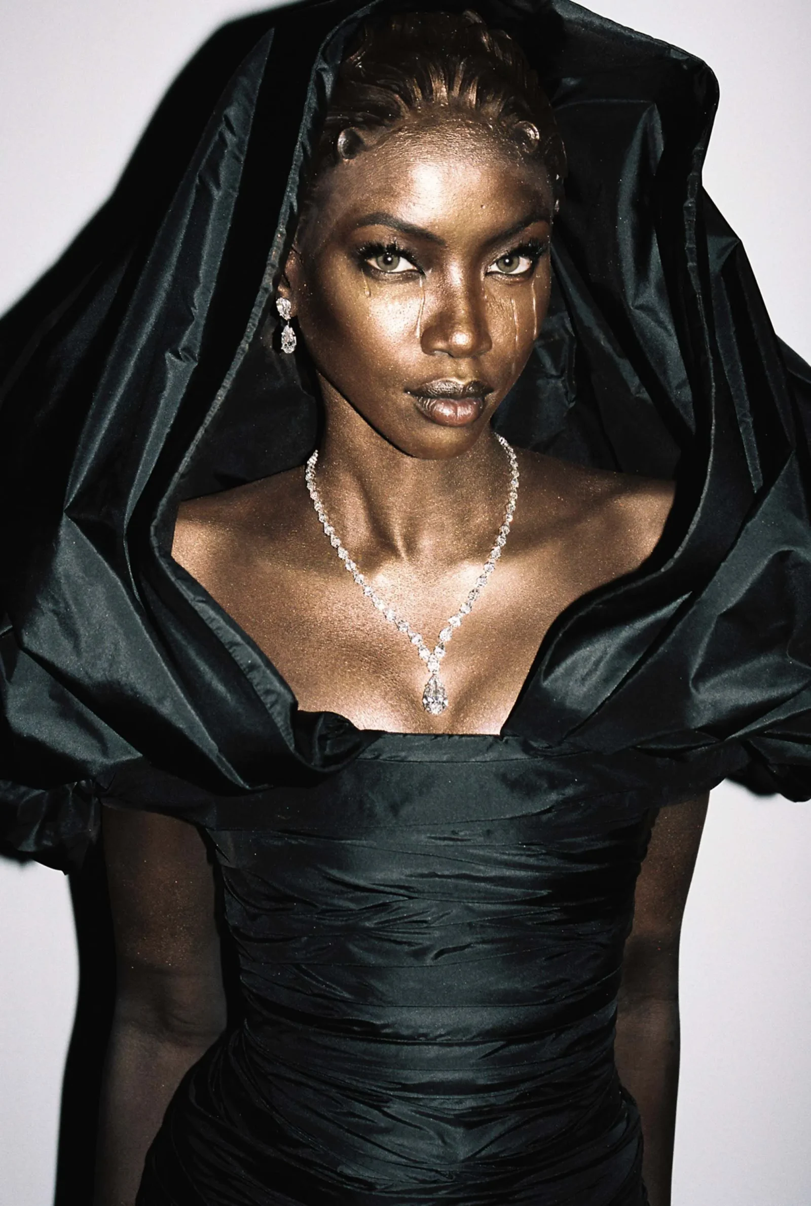 Woman in an elegant black satin gown with a dramatic draped neckline and veil, wearing diamond jewelry and looking at the camera.