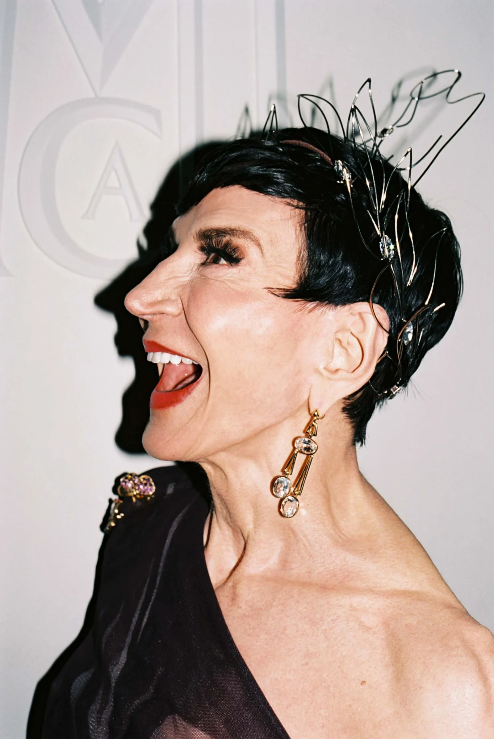Profile view of a laughing woman with short black hair, wearing a metallic, sculptural headpiece and large jeweled earrings.