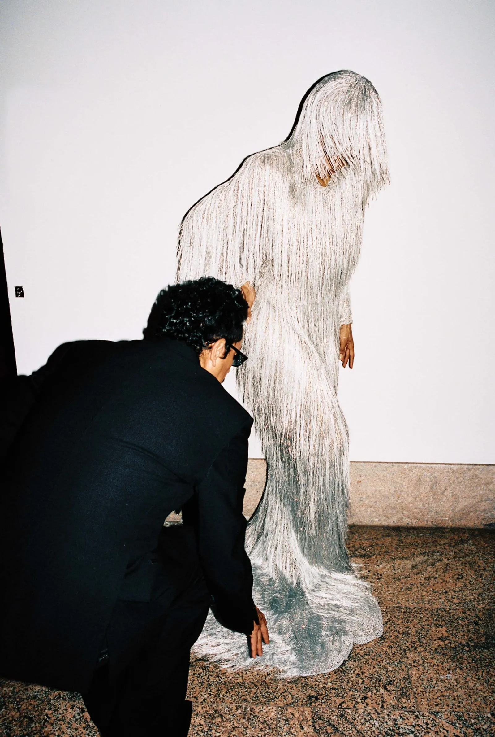 Man in a dark suit kneels and touches the base of a tall, silver-white fringed dress sculpture against a plain wall.
