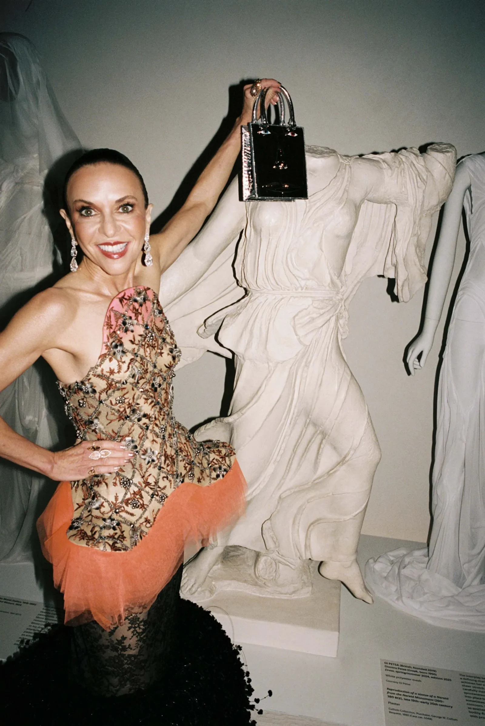 Smiling woman in an ornate floral gown holds up a shiny black handbag beside a white statue.