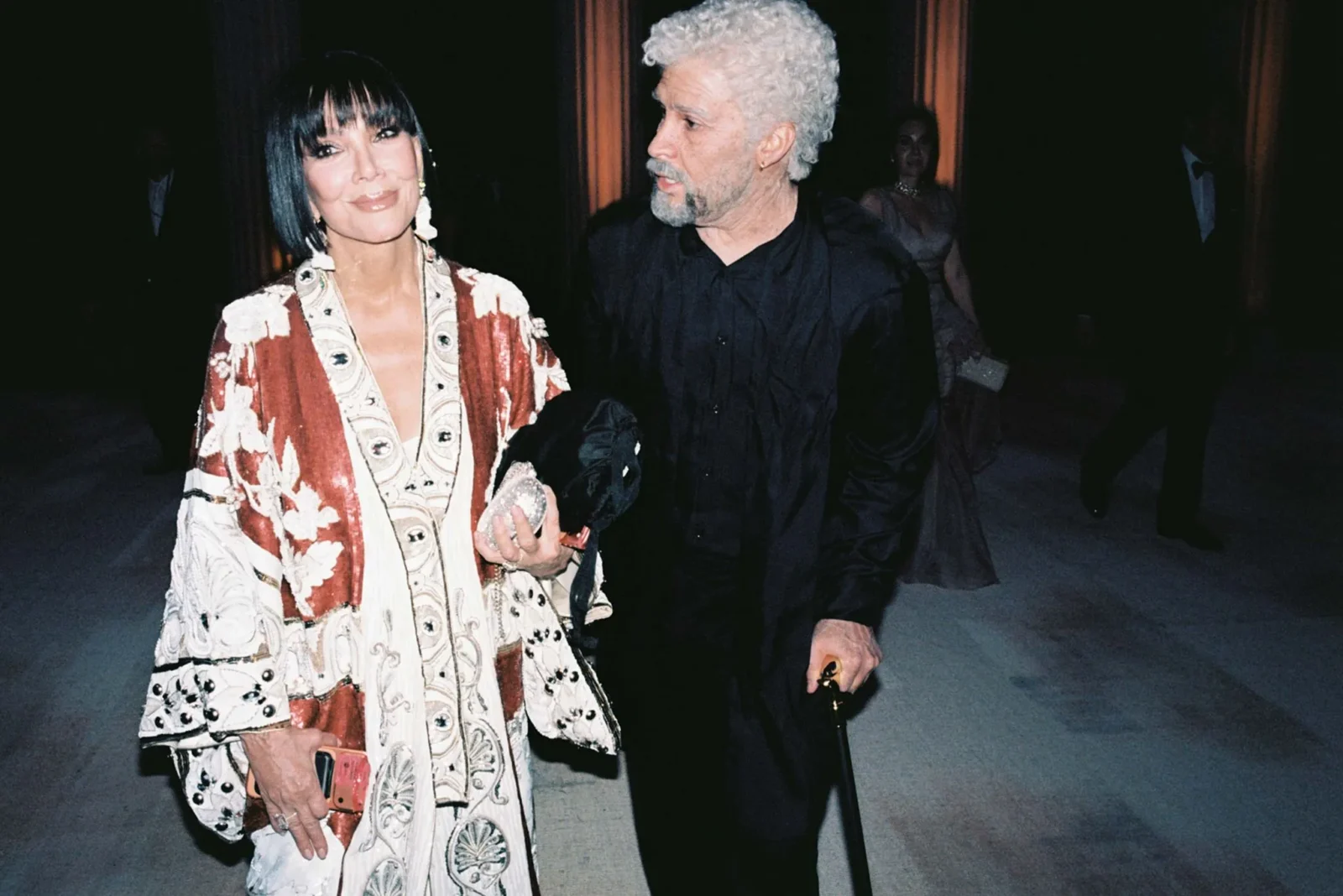 Two elegantly dressed adults walk together at a formal event; woman in a flowing patterned cream‑red dress, man in a black suit behind her.