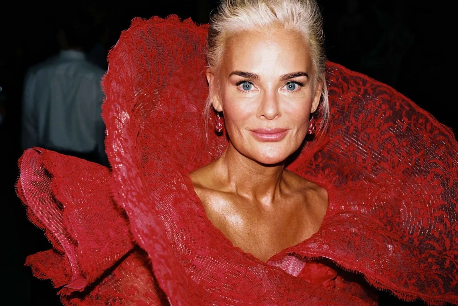 Close-up of a woman with blue eyes and blonde hair wearing a bold red lace dress with dramatic ruffles around the shoulders.