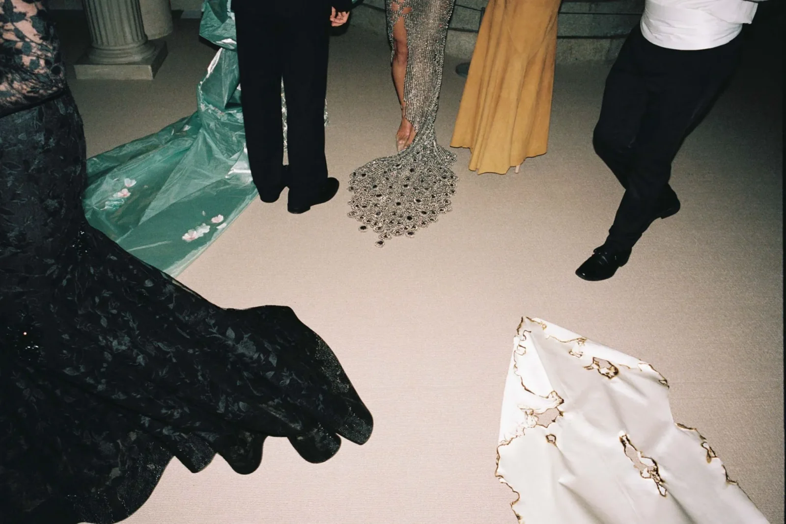 People in formal attire standing on a beige carpet; long gowns and tuxedos with a beaded train visible at the center.
