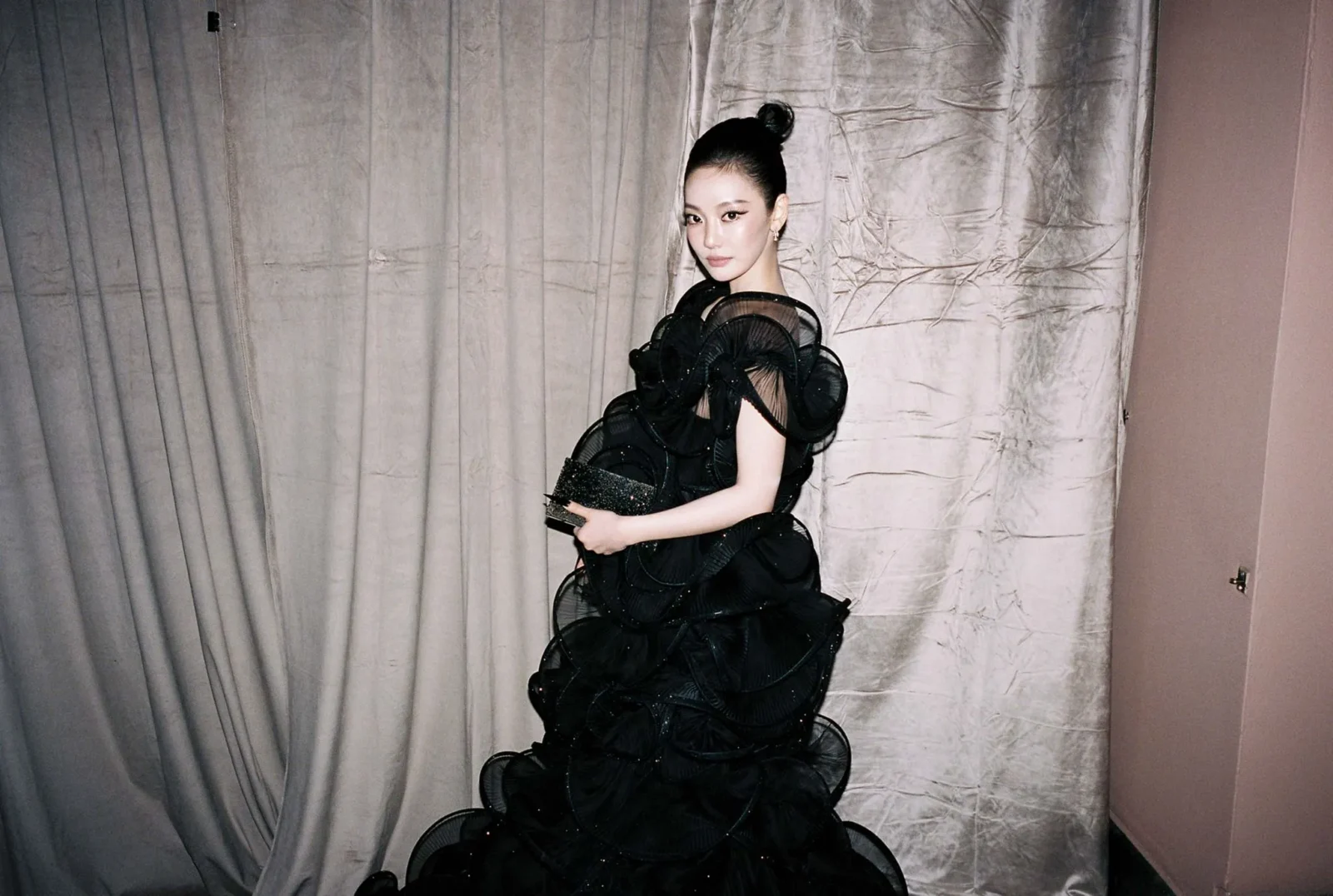 Woman in an elaborate black ruffled gown posing against a light-colored curtain backdrop, looking at the camera.