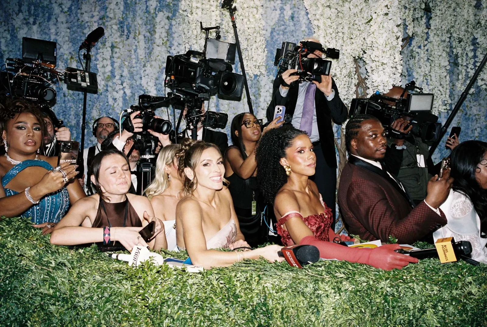 Crowded red-carpet scene with photographers and reporters aiming cameras at guests against a wall of white flowers and greenery.