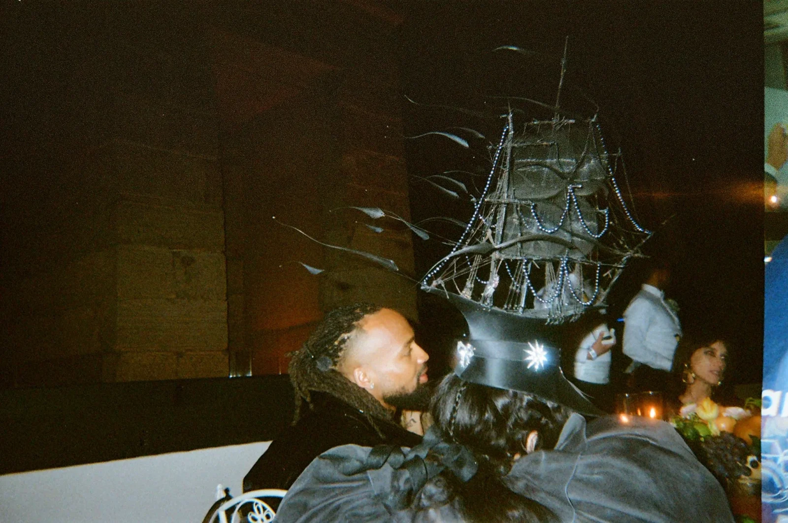 Nighttime party scene with guests in black outfits; a performer wears an elaborate beaded headpiece with lights.