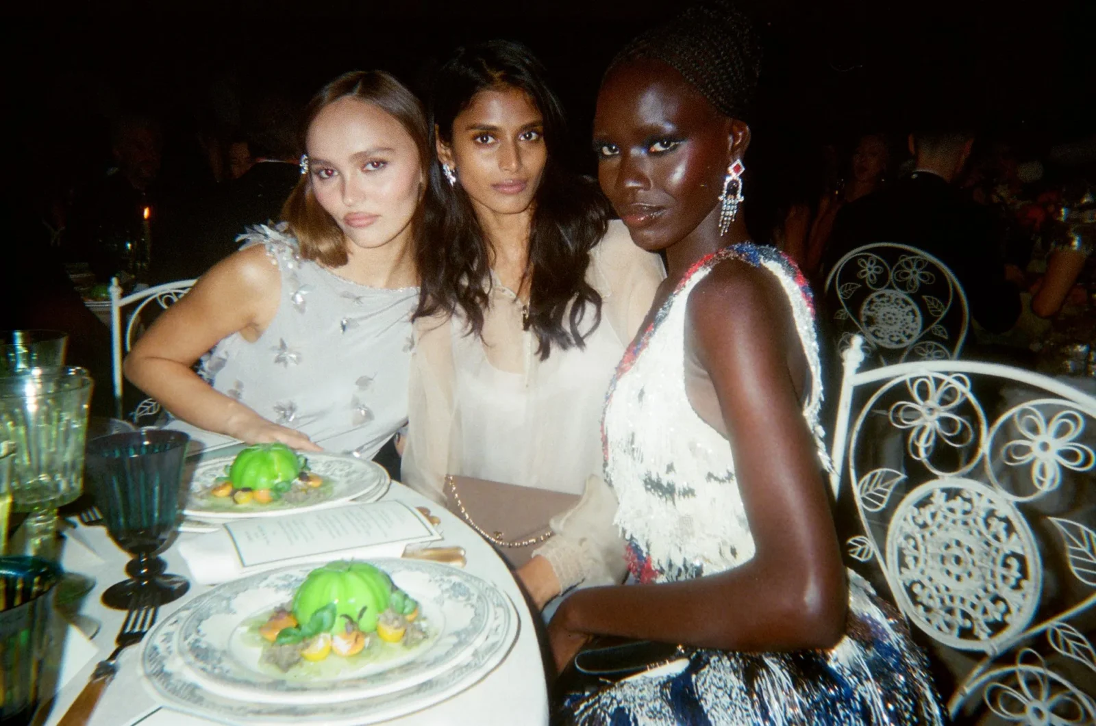 Three women in evening dresses pose at a dinner table with plates and green desserts on the plate area.